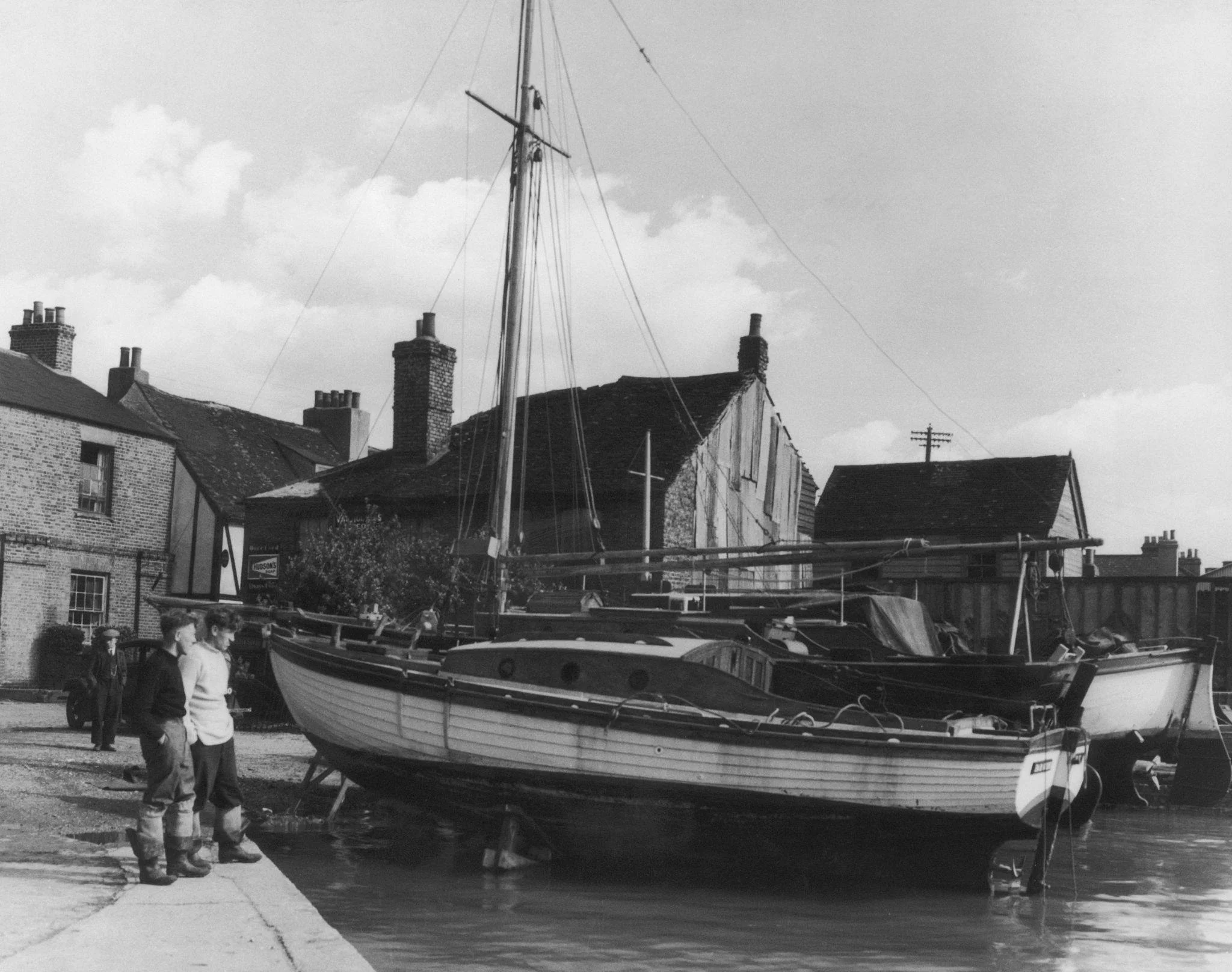 A black-and-white photo of a boat on land, supported by a trailer, with buildings and a boat mast in the background, and a few people standing nearby.