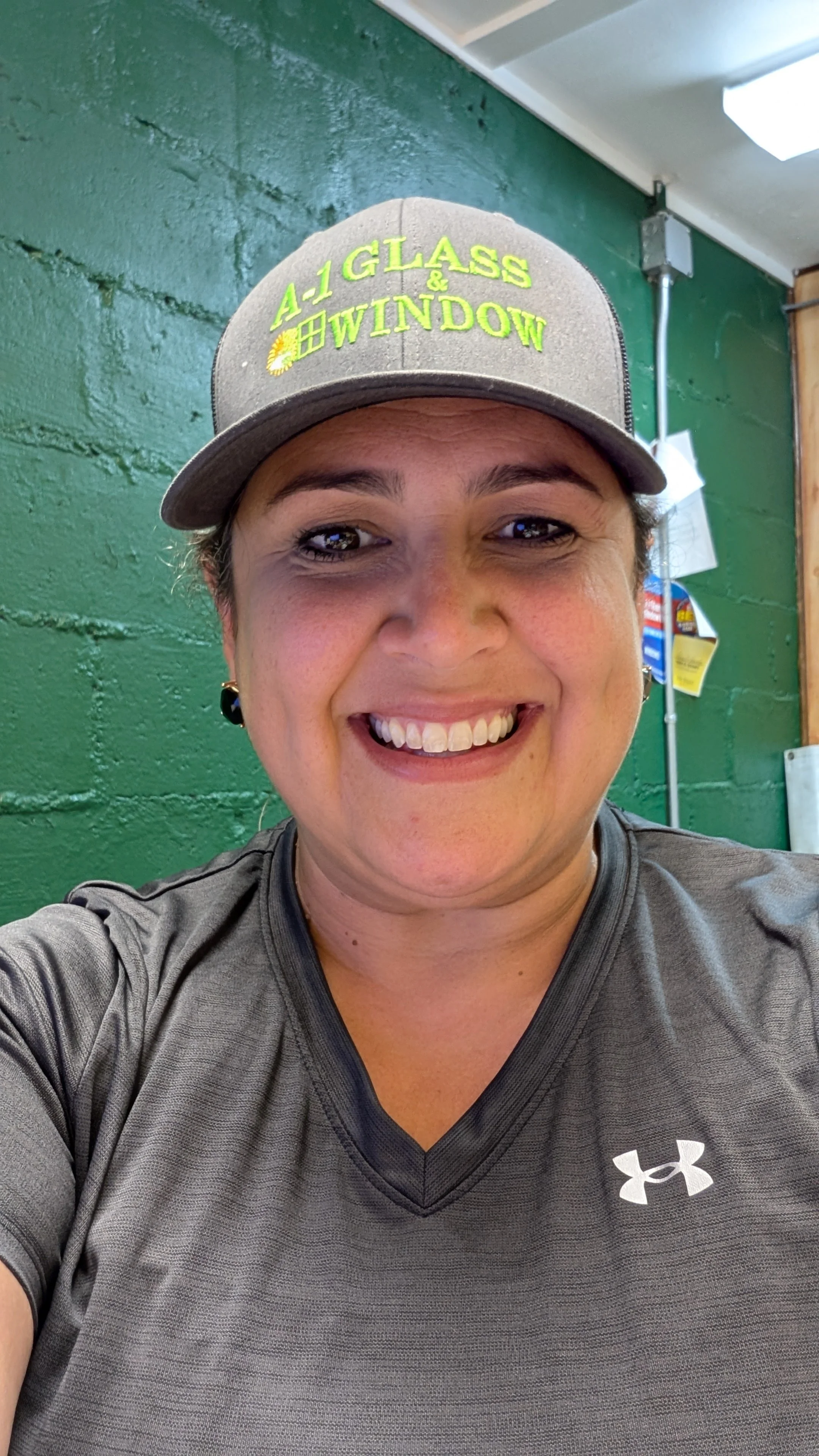 A woman smiling in a gym wearing a gray Under Armour shirt and a gray cap that says "A-1 Glass & Window".