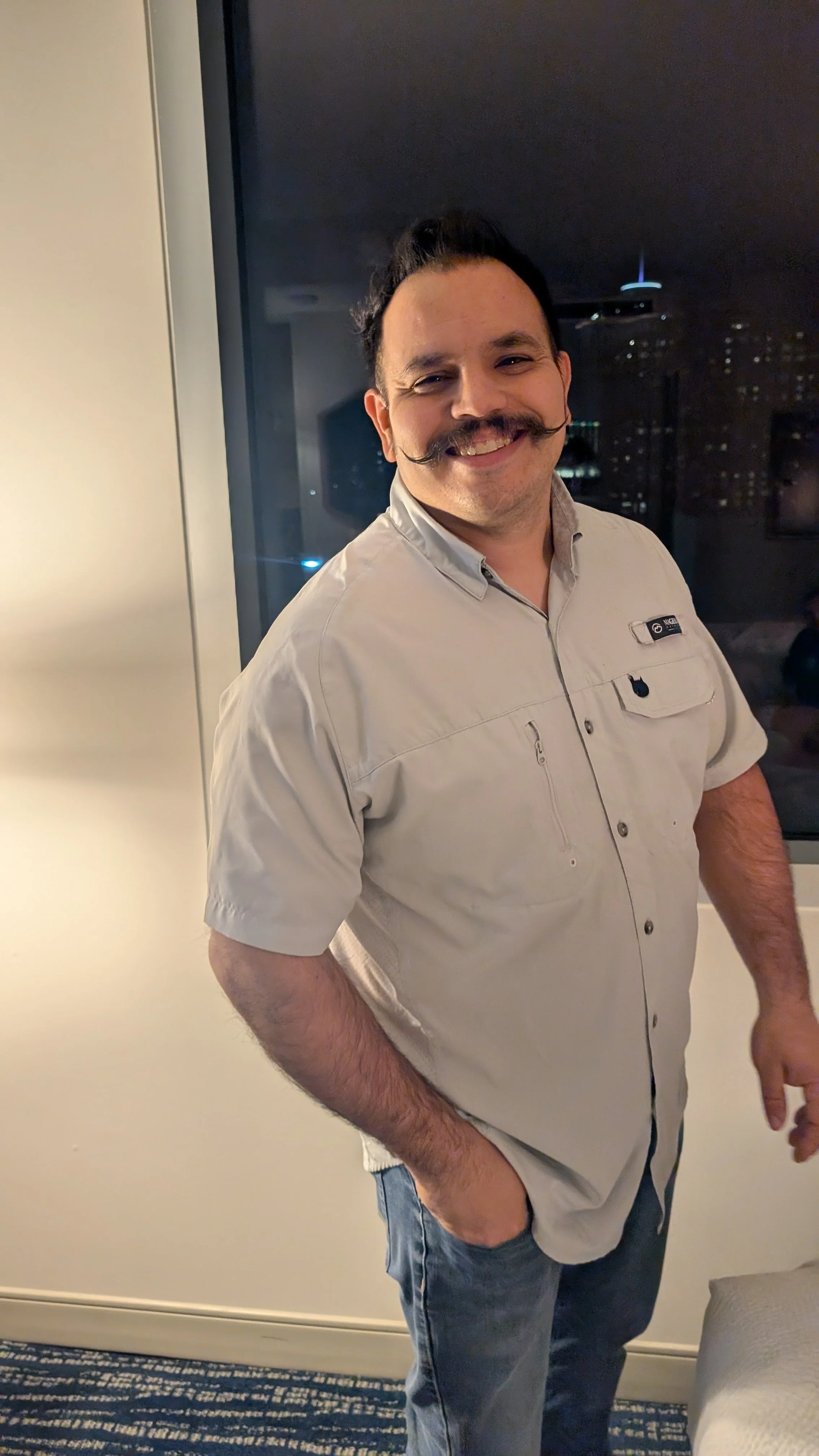 A man with a styled dark mustache, wearing a beige button-up shirt and jeans, smiling and posing indoors at night.