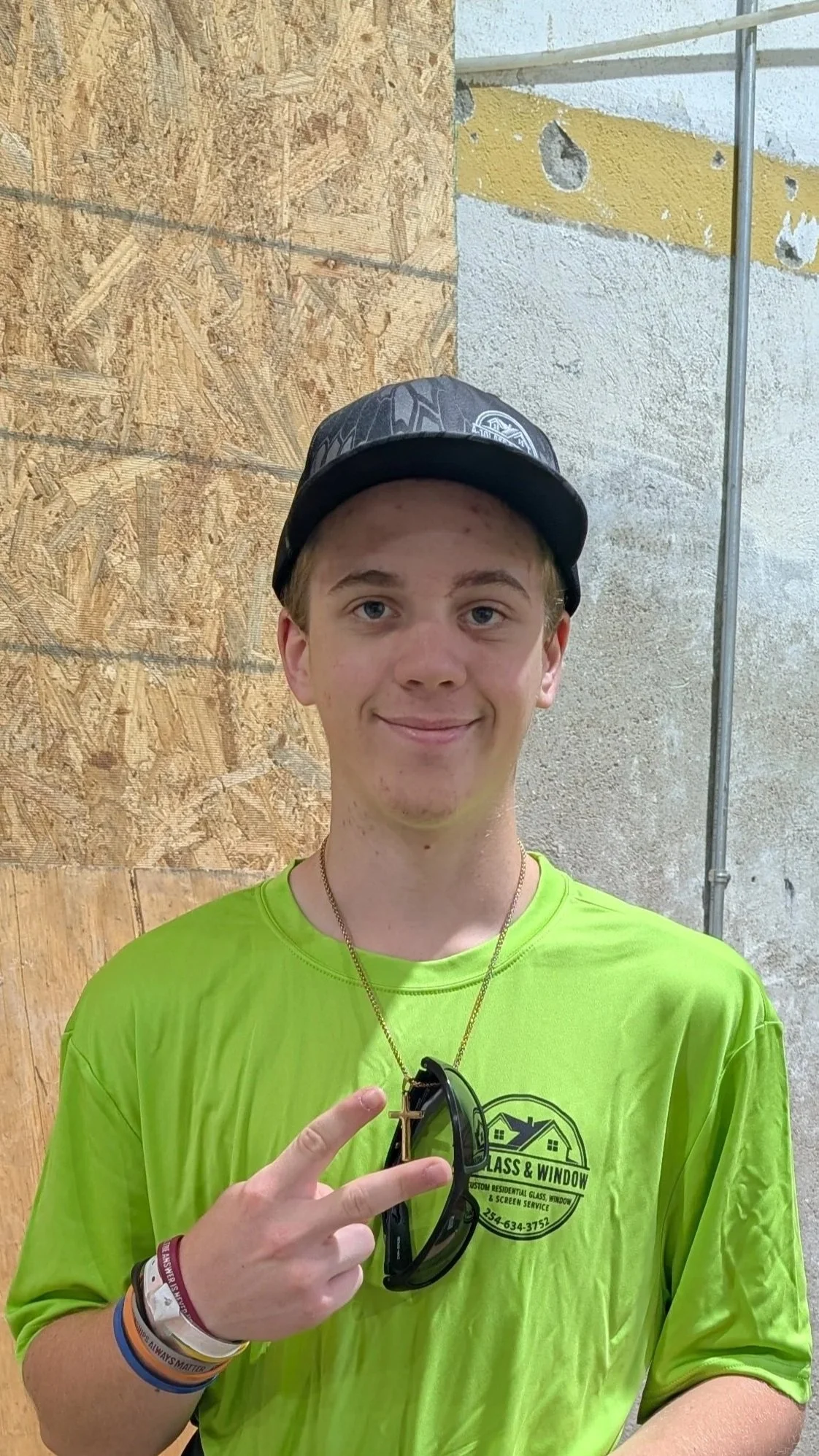 A smiling young man with light skin, blond hair, and wearing a black cap and bright green t-shirt. He is holding sunglasses and pointing with his fingers, wearing multiple wristbands, one of which reads 'Always Matter.' The background features a wood and concrete wall, with metal piping above.