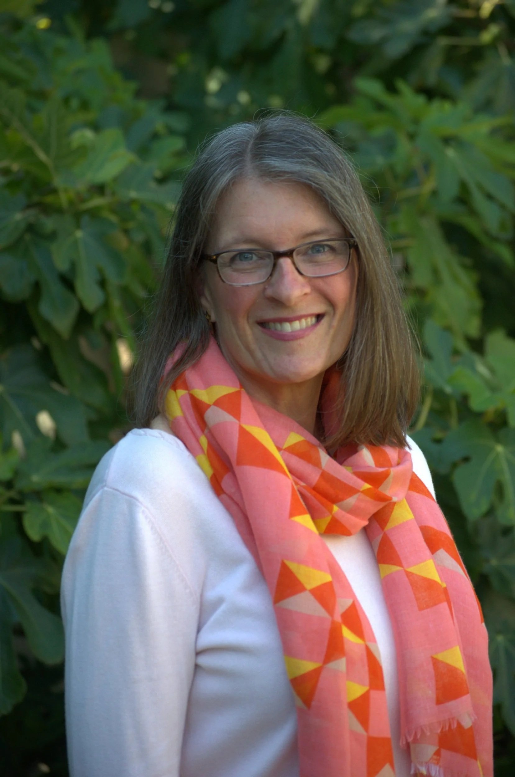 A woman with long brown hair, wearing glasses, a white top, and an orange and yellow patterned scarf, smiling outdoors with green foliage in the background.