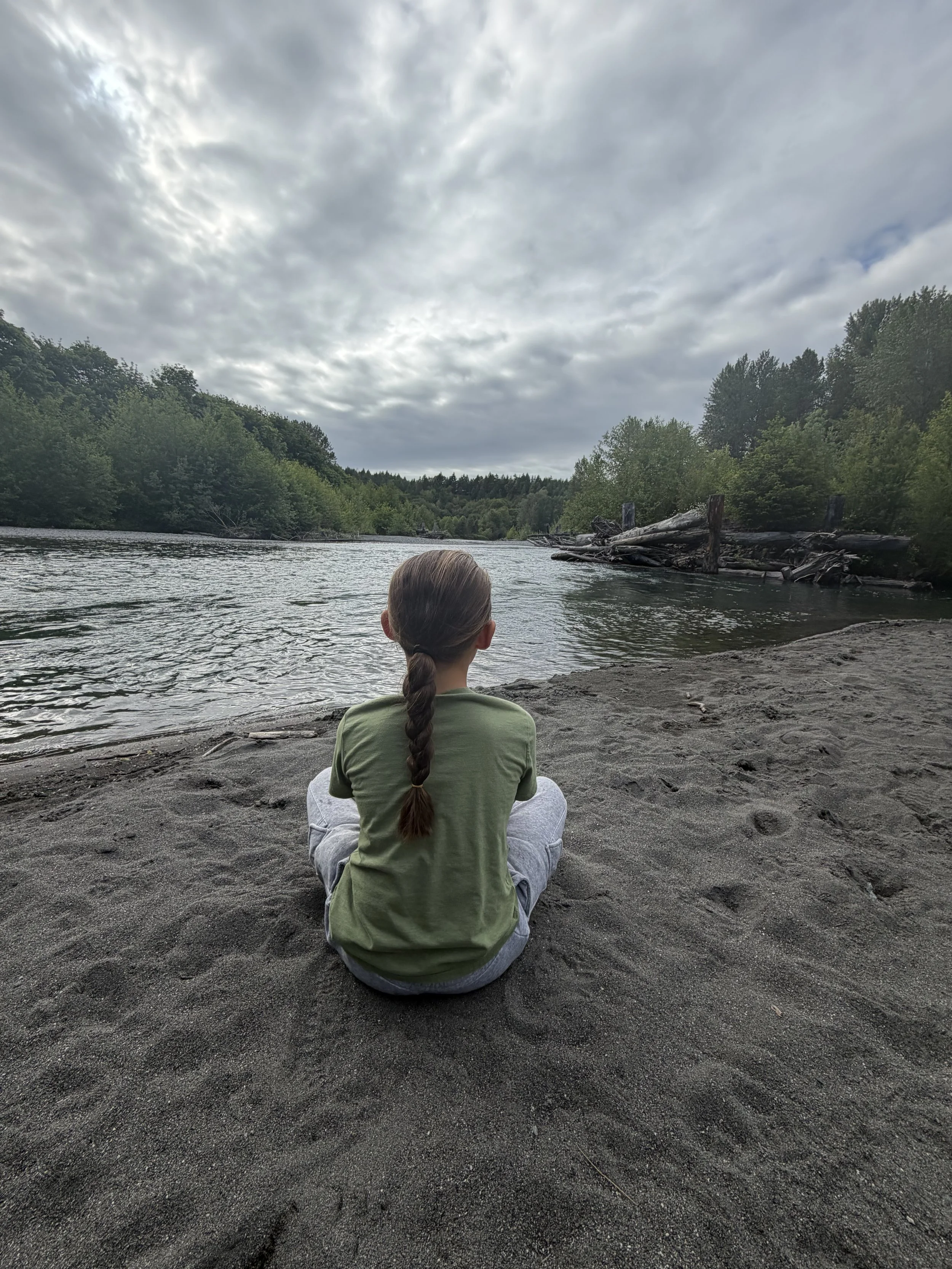 A person with long brown hair in a braid, wearing a green shirt, is sitting cross-legged on a sandy beach near a river, facing away and looking towards the water and cloudy sky.