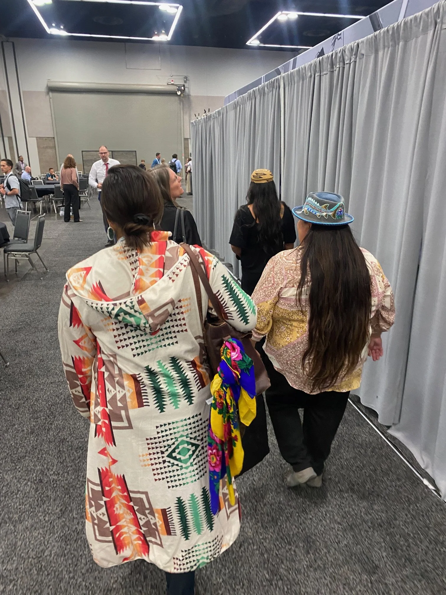 People standing in line at an indoor event, with some wearing traditional Indigenous clothing and accessories, enclosed by gray curtains.