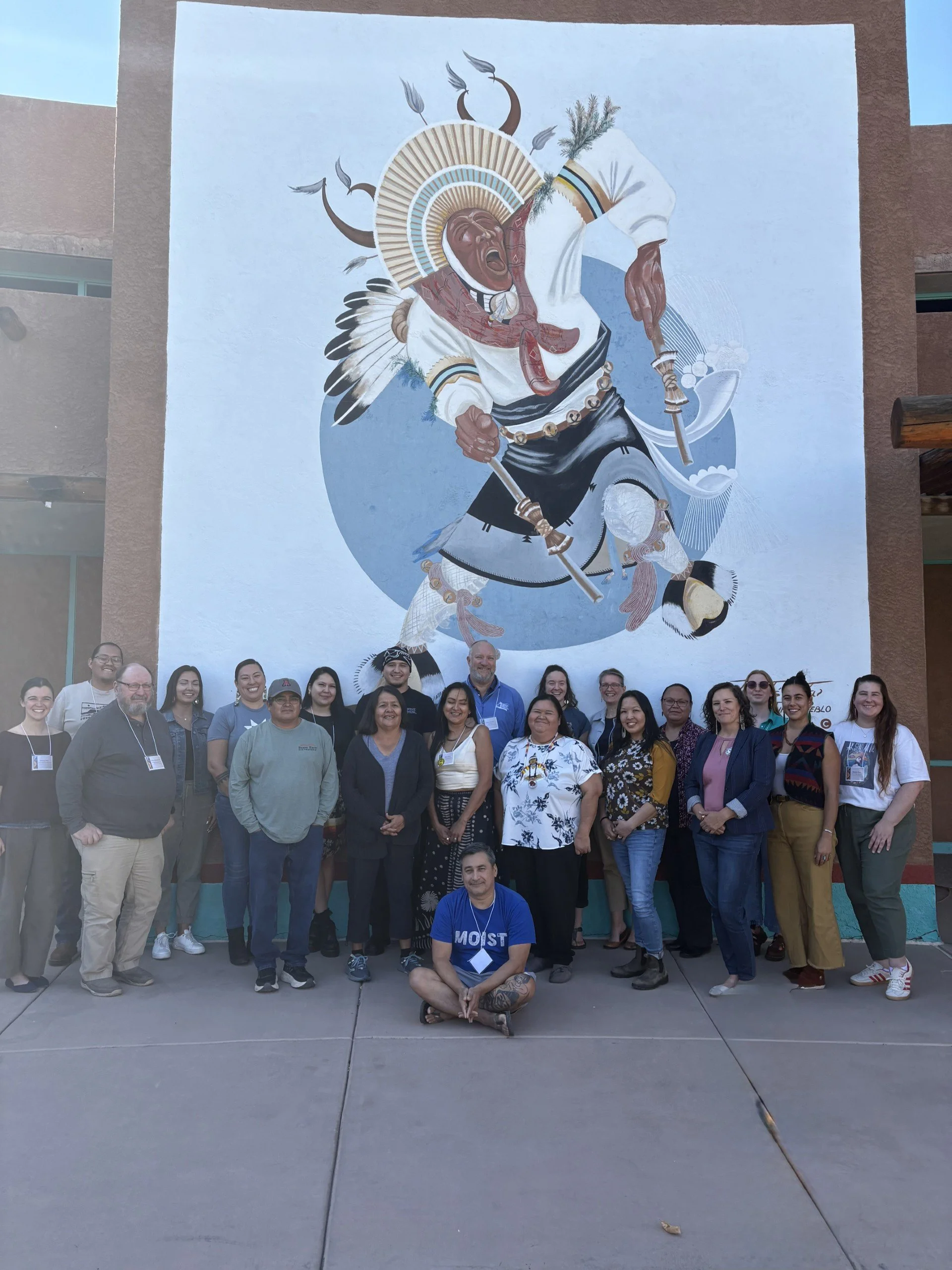 A group of diverse people standing outside in front of a large mural of a Native American warrior. The mural features the warrior wearing a feathered headdress, holding a staff, and dressed in traditional attire. Some people are smiling and posing for a group photo, with one person sitting cross-legged on the ground in front.