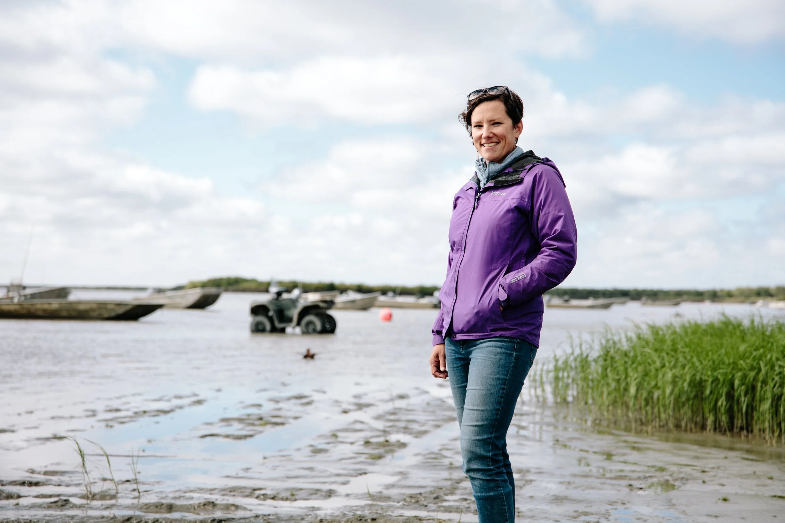 Woman standing on the shore near a body of water with boats and an all-terrain vehicle in the background, wearing a purple jacket and jeans.