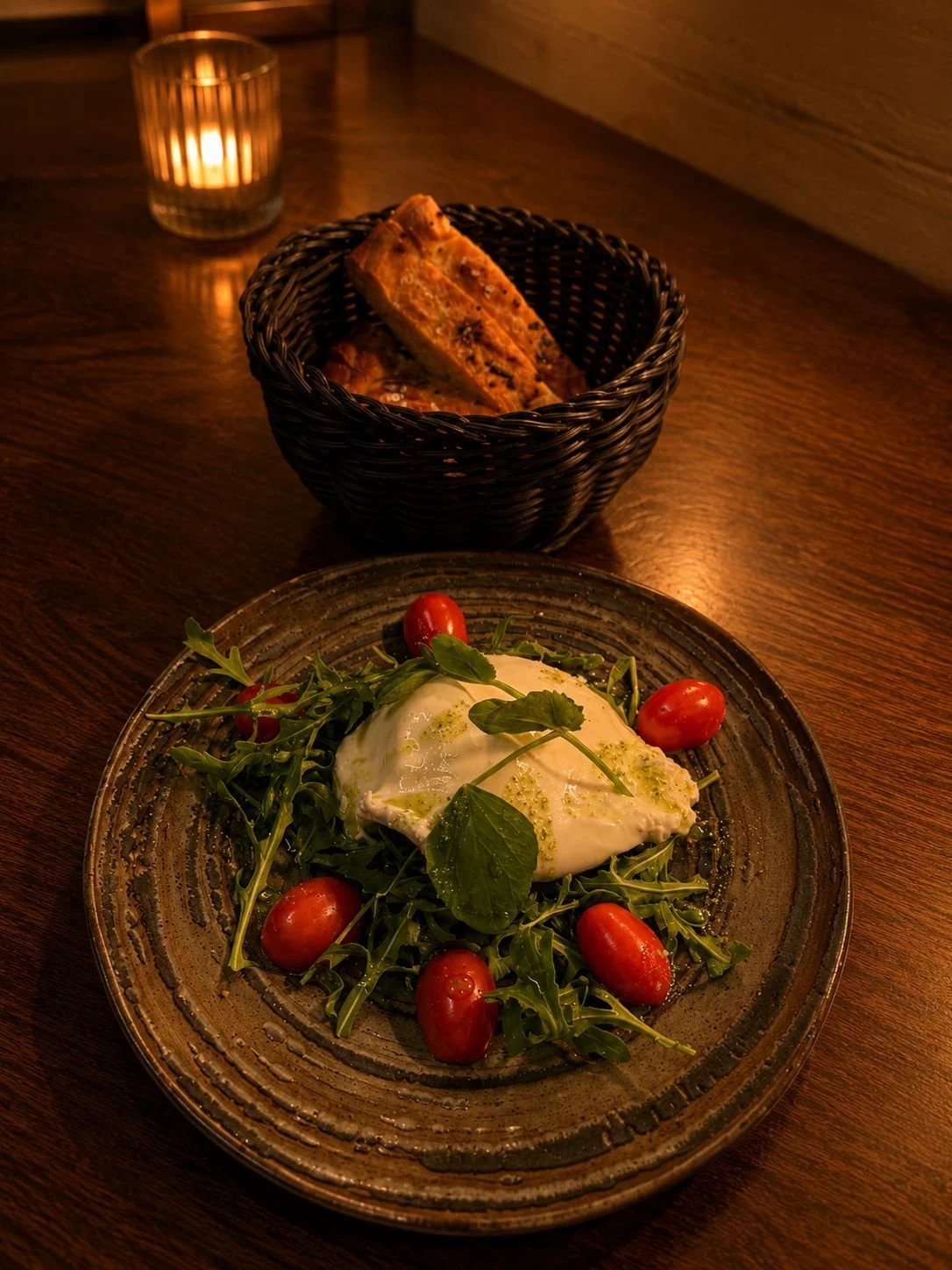 A plate of salad with cherry tomatoes, greens, and a poached egg, accompanied by slices of baguette bread in a basket, and a lit candle in the background.