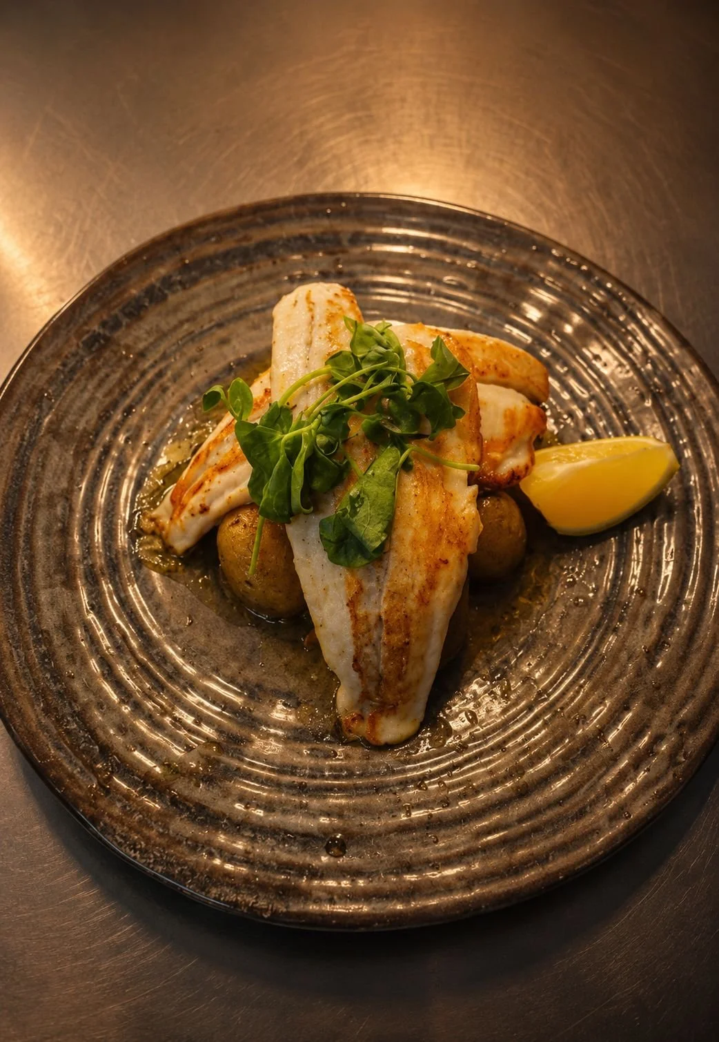 Seared fish fillet on a dark ceramic plate with a lemon wedge, microgreens, and small round potatoes.
