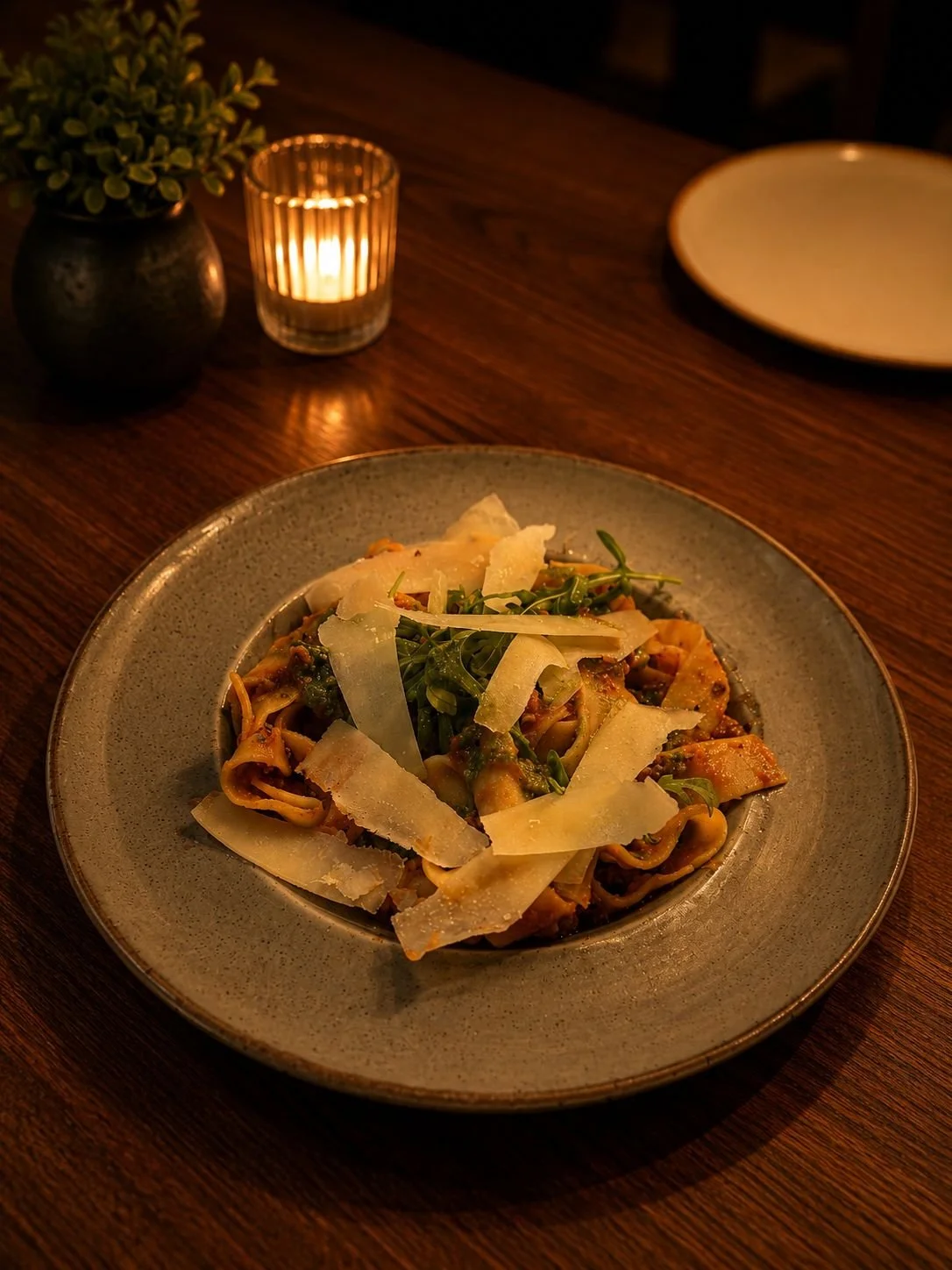 Plate of pasta topped with parmesan cheese, garnished with greens, on a wooden table illuminated by candlelight