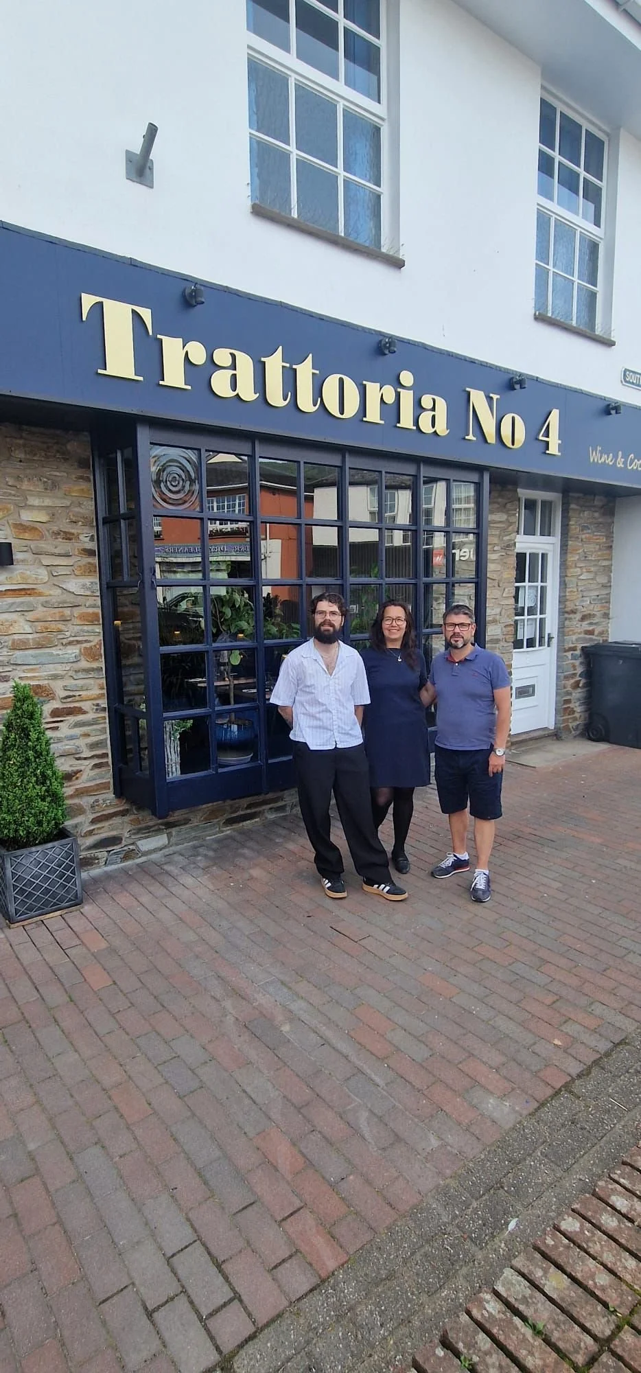 Three people standing in front of a restaurant called 'Trattoria No 4,' with brick sidewalk and a storefront window with plants inside.
