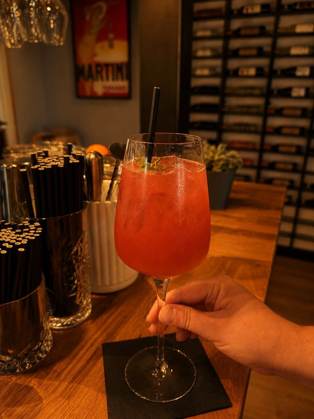A hand holding a tall glass of pink cocktail with ice, garnished with a mint sprig and a straw, on a black napkin at a wooden bar counter.