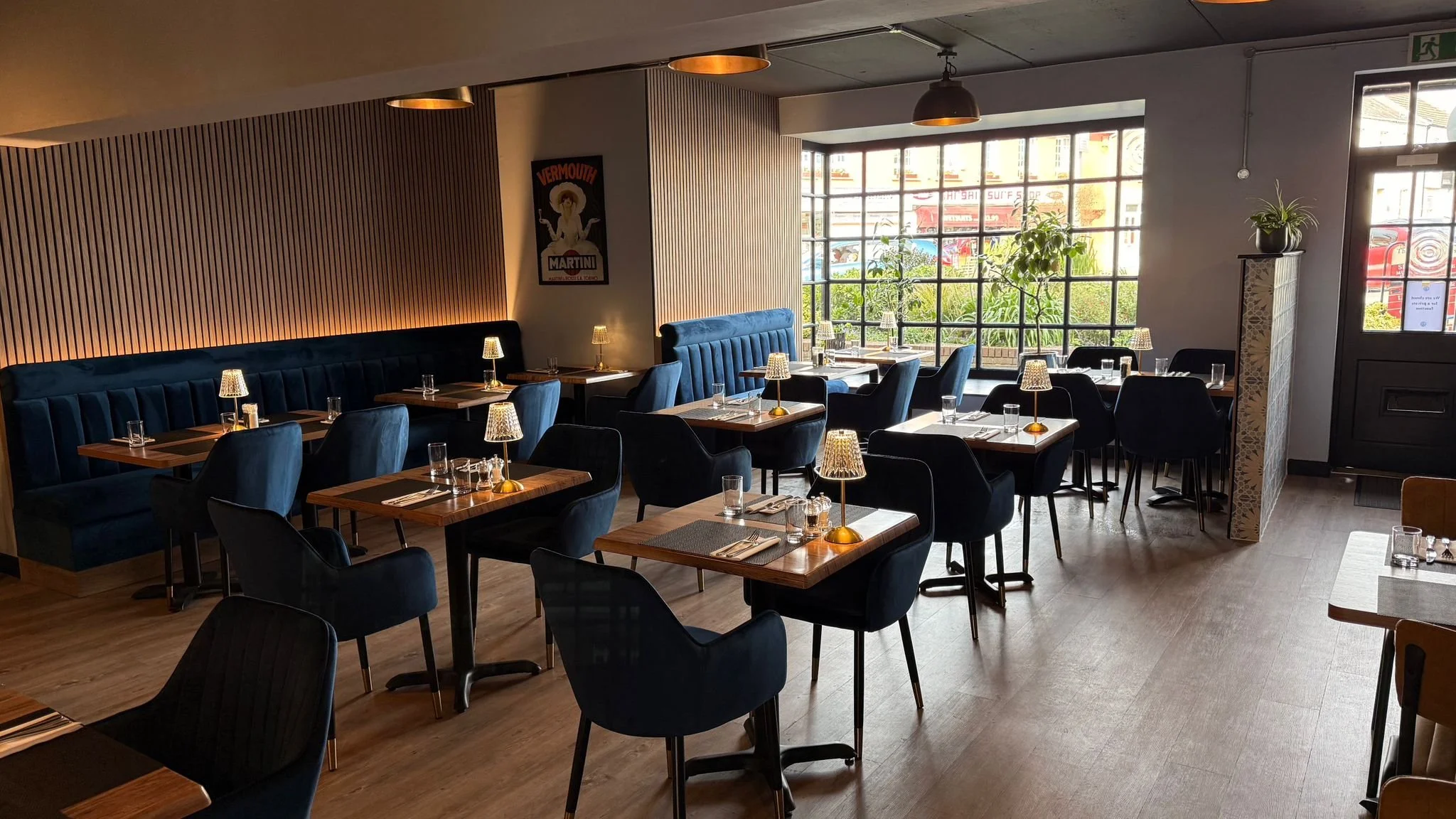 Interior of a modern restaurant with dark blue chairs and wooden tables, illuminated by small table lamps, large windows with natural light, and decorative wall art.