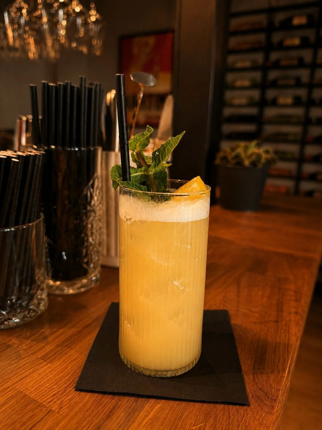 A tall glass of a yellowish cocktail garnished with a lemon wedge and mint leaves, with two black straws, on a black napkin on a wooden bar counter.