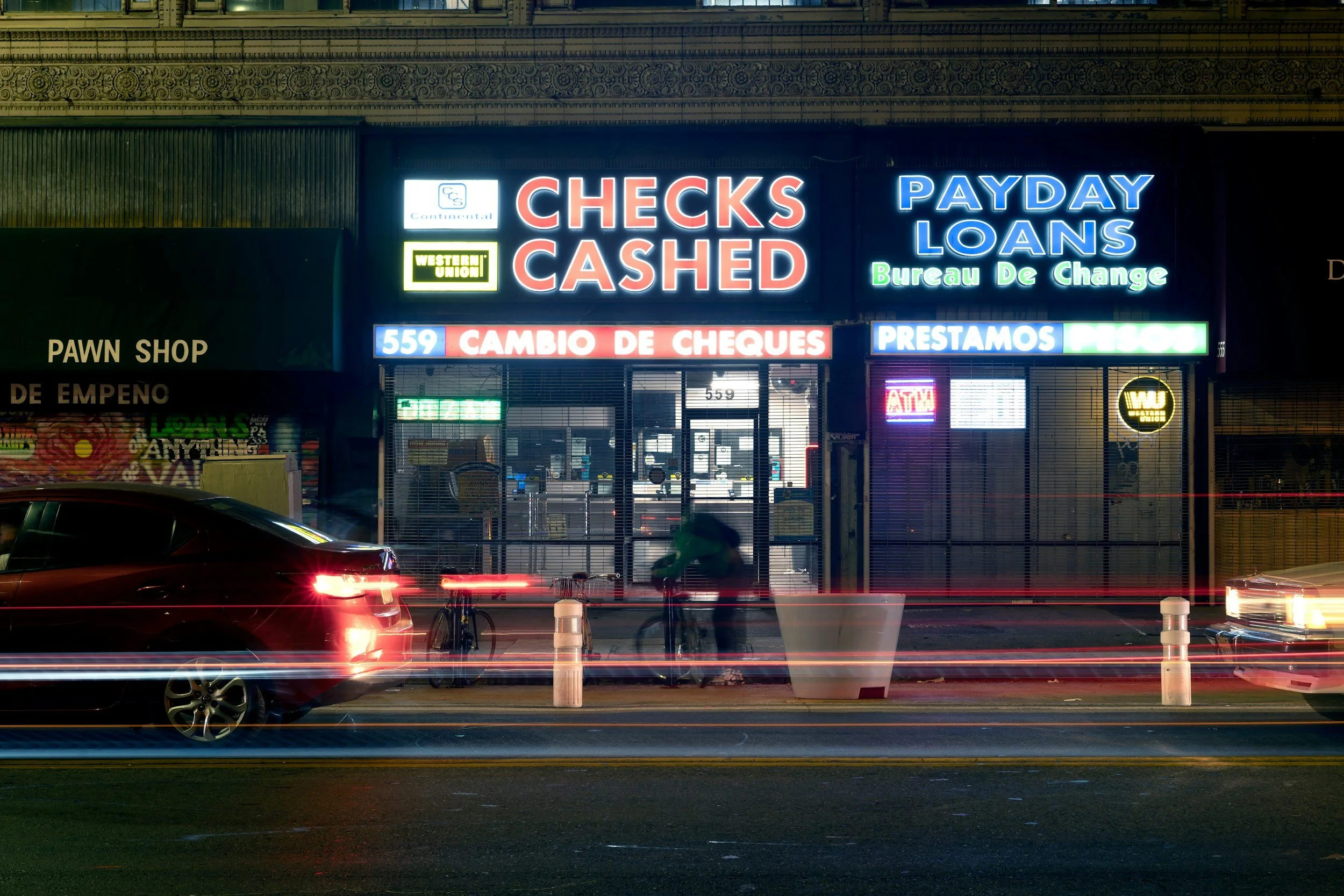 Neon signs on a storefront advertising checks cashed, payday loans, and currency exchange service at night. There are cars and a bicycle in front of the shop, with streaks of car lights indicating long exposure.