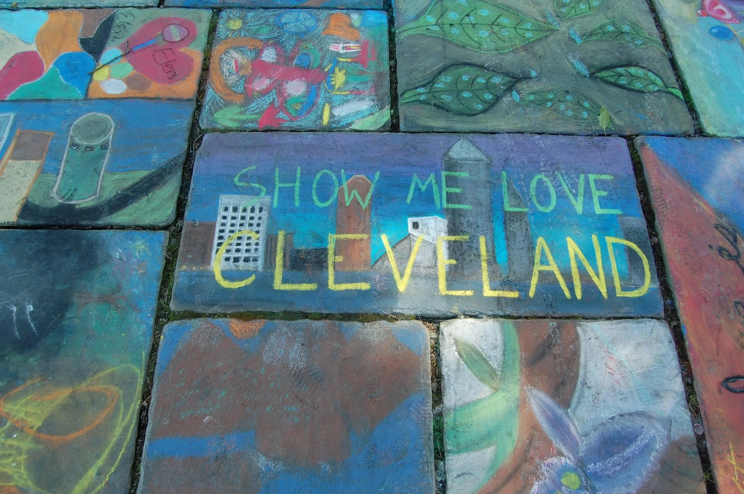 Colorful sidewalk chalk paintings on stones, including a message that says 'SHOW ME LOVE CLEVELAND' with illustrations of buildings and cityscape.