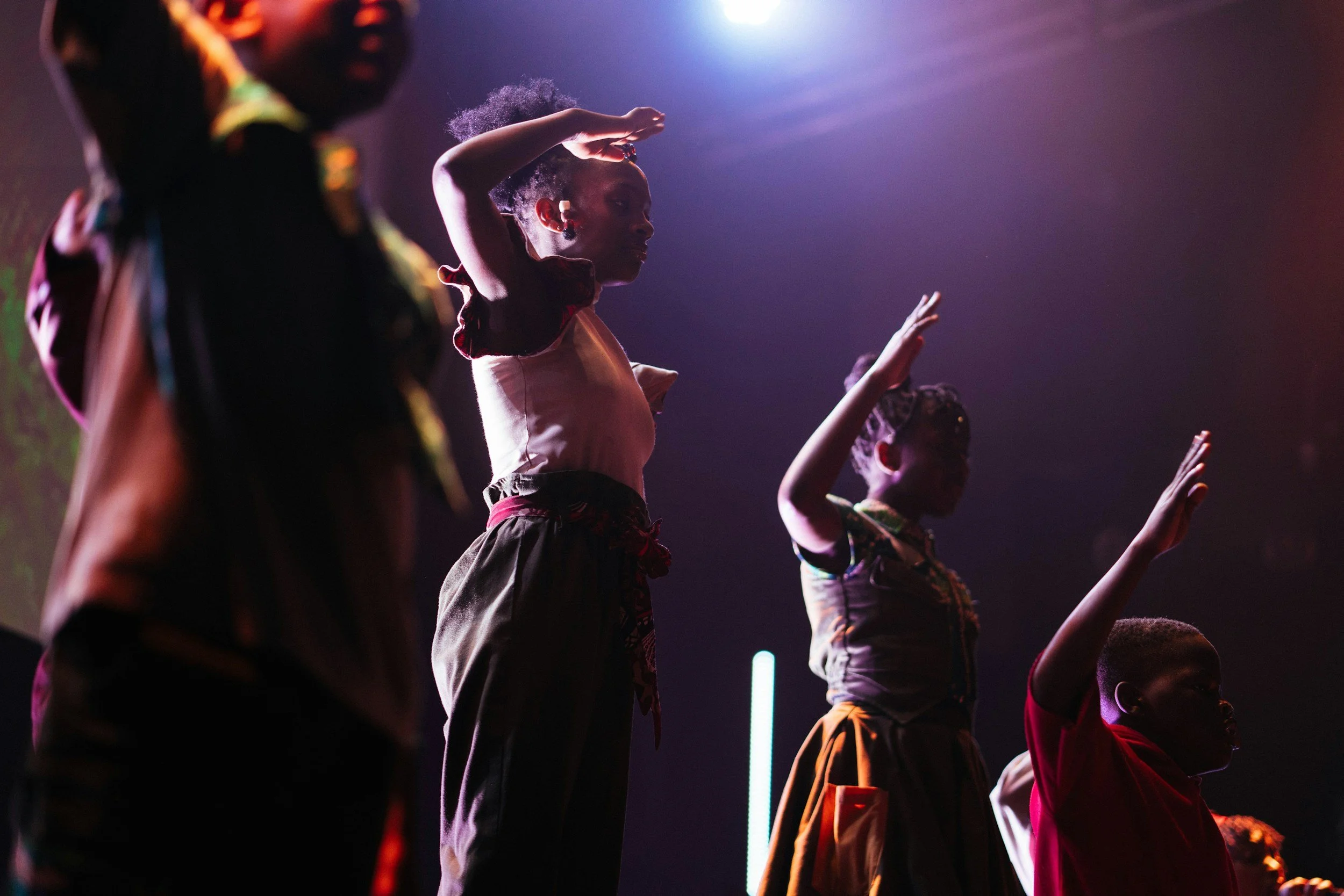 Young girls dressed in colorful traditional clothing dancing on stage with vibrant lighting.