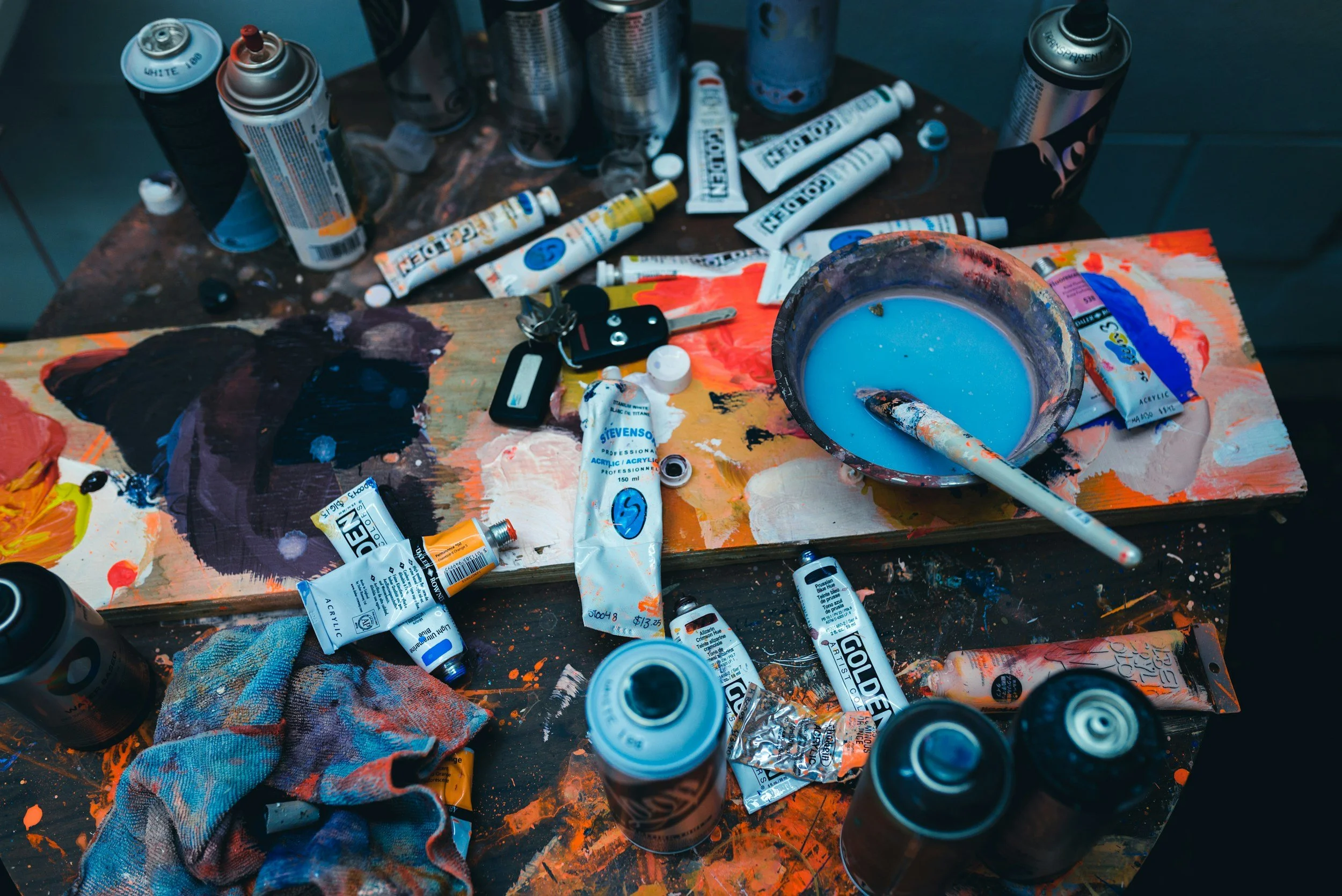 A cluttered artist's workspace with open and closed paint tubes, spray paint cans, a bowl of blue paint with a painted brush, and a dirty cloth on a splattered table.