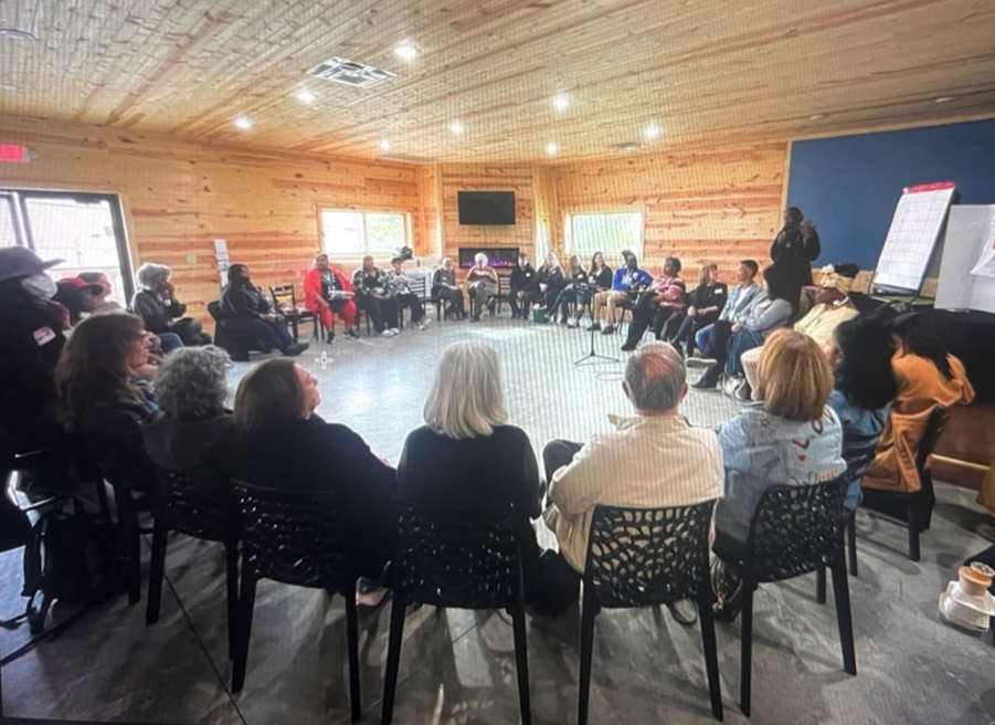 A group of people seated in a large, circular arrangement in a wood-paneled room, participating in a meeting or workshop, with a speaker standing and addressing the group.