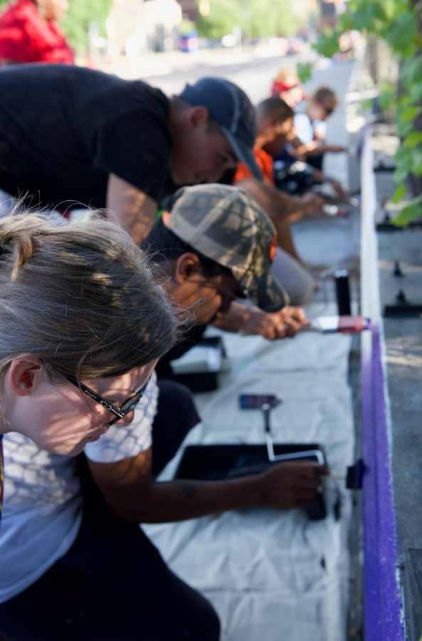 People painting a mural outdoors on a purple wall with brushes and paint.