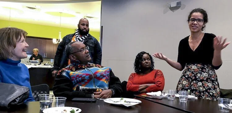 A woman speaking to a group of four people seated at a table, with a plate and glasses, in a room with a kitchen area in the background.