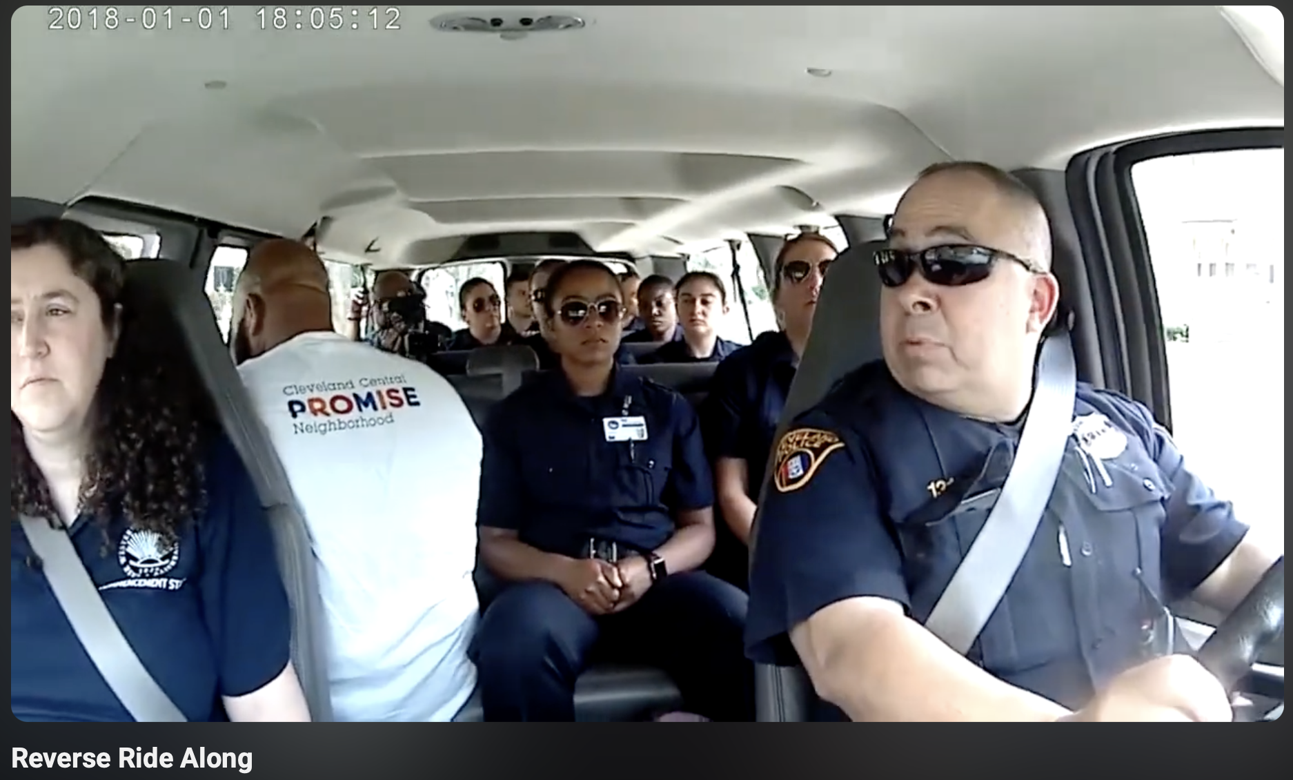 Inside a vehicle, five police officers and one woman are seated, with officers wearing uniforms and sunglasses. The front driver is a male officer with a shaved head and sunglasses. Behind him, a woman with dark hair and sunglasses sits next to a woman wearing a white shirt that has 'Promise Neighborhood' written on the back. Further back, several individuals, including a woman with a camera, are visible, and the vehicle appears to be in daytime with sunlight outside.