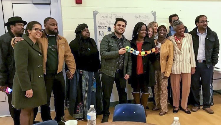 Group of diverse adults standing in a classroom, smiling, holding a paper chain, with posters and a whiteboard in the background.