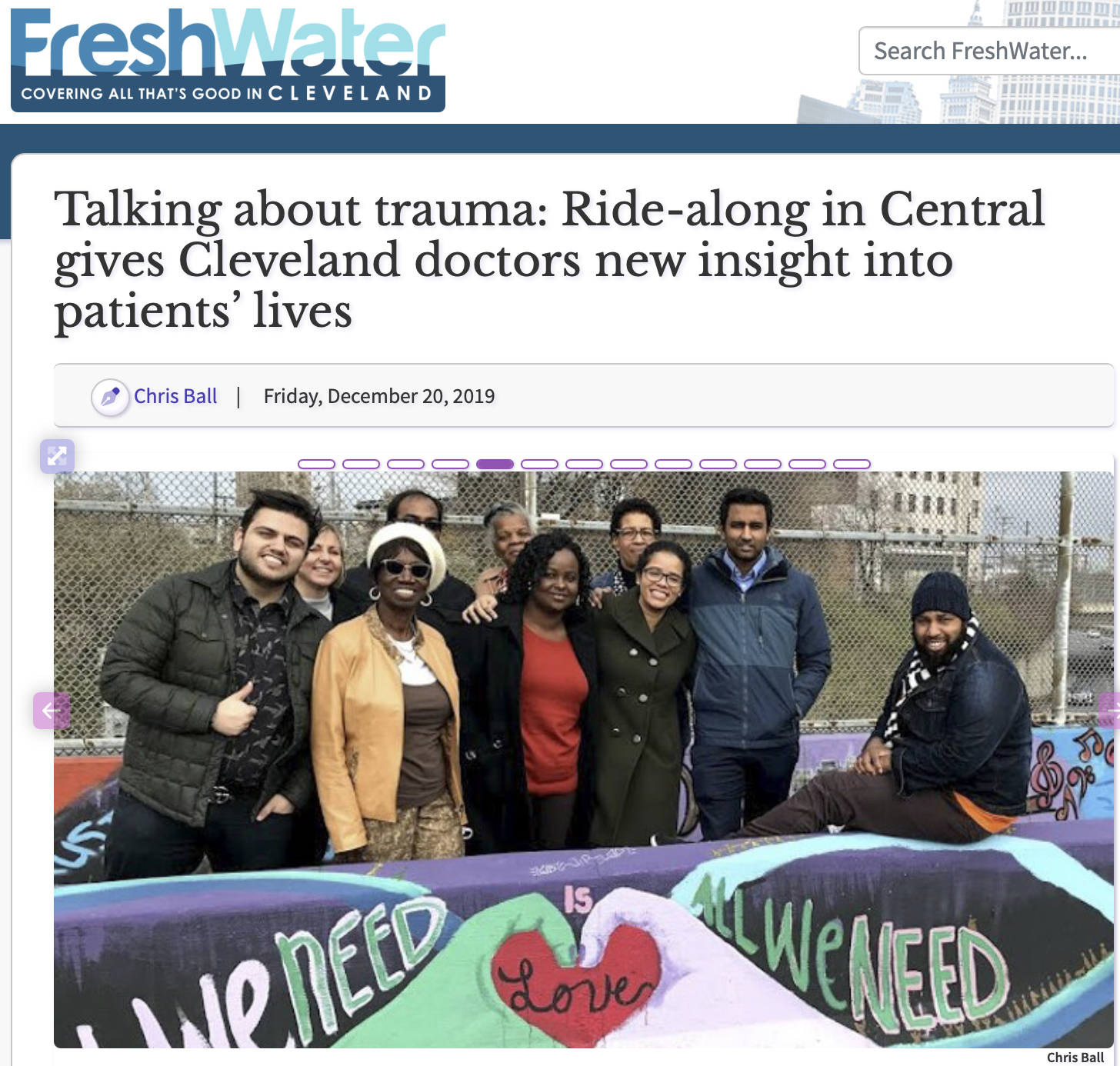 Group of diverse people standing outdoors behind a colorful banner with words about needs and love, in front of a chain-link fence and urban buildings.