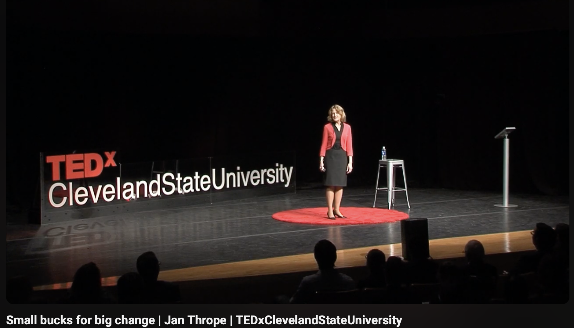 A woman standing on stage at TEDxClevelandStateUniversity. She is wearing a red blazer and black dress, next to a stool with a water bottle. The stage has a large sign reading 'TEDx Cleveland State University' and a black podium.