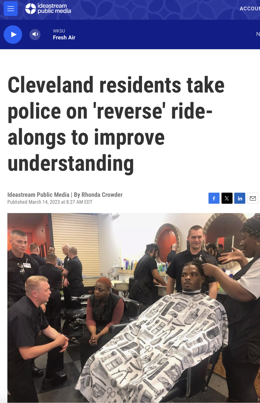 Police officers and a barber in a barbershop, with a man sitting in a barber's chair getting a haircut, surrounded by other officers and staff.