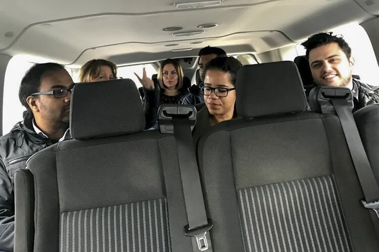 Group of six people sitting in the back of a vehicle, some making peace signs and smiling.