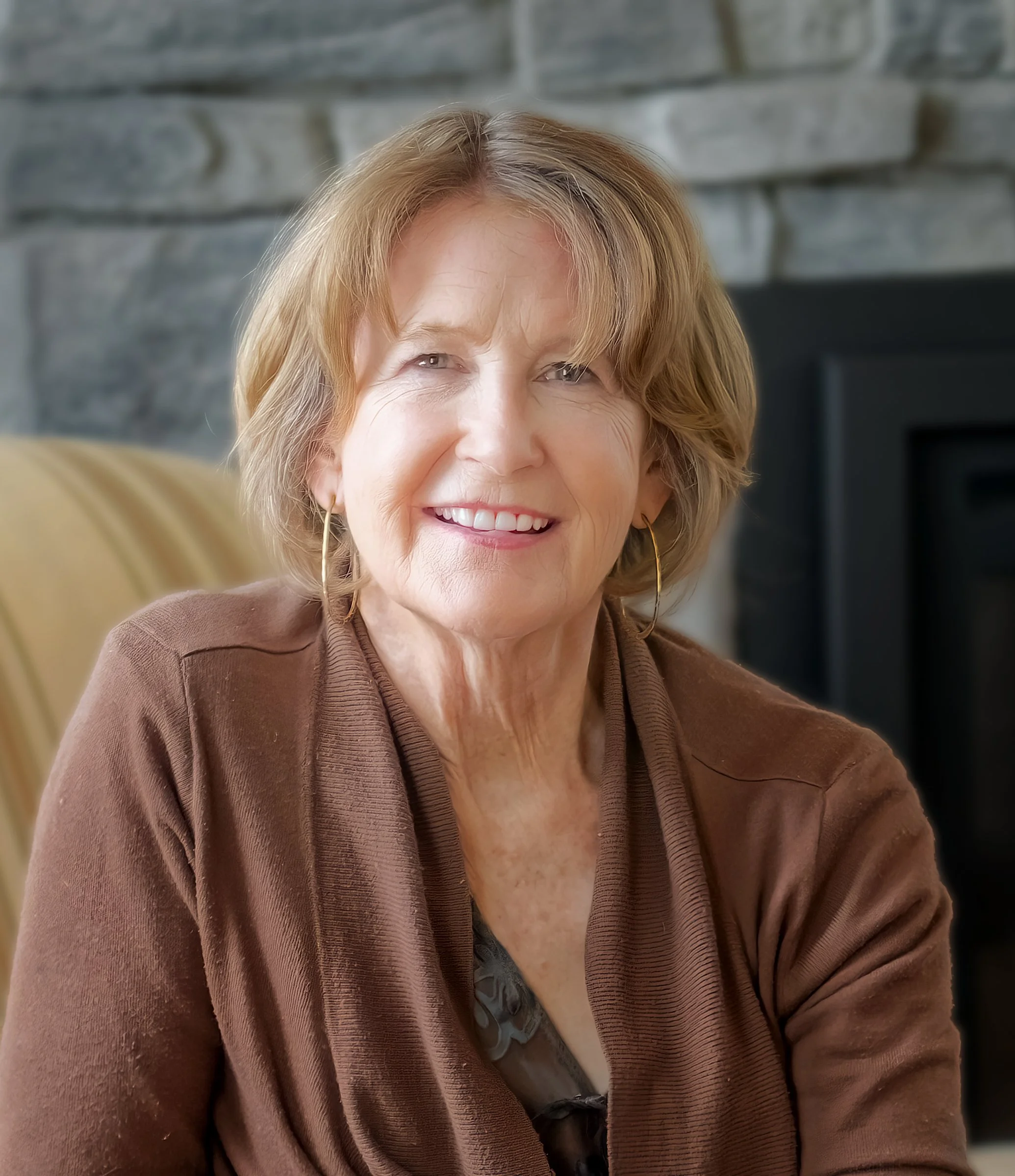 Close-up of an elderly woman with short blonde hair, smiling, wearing hoop earrings and a brown cardigan, in front of a stone wall.