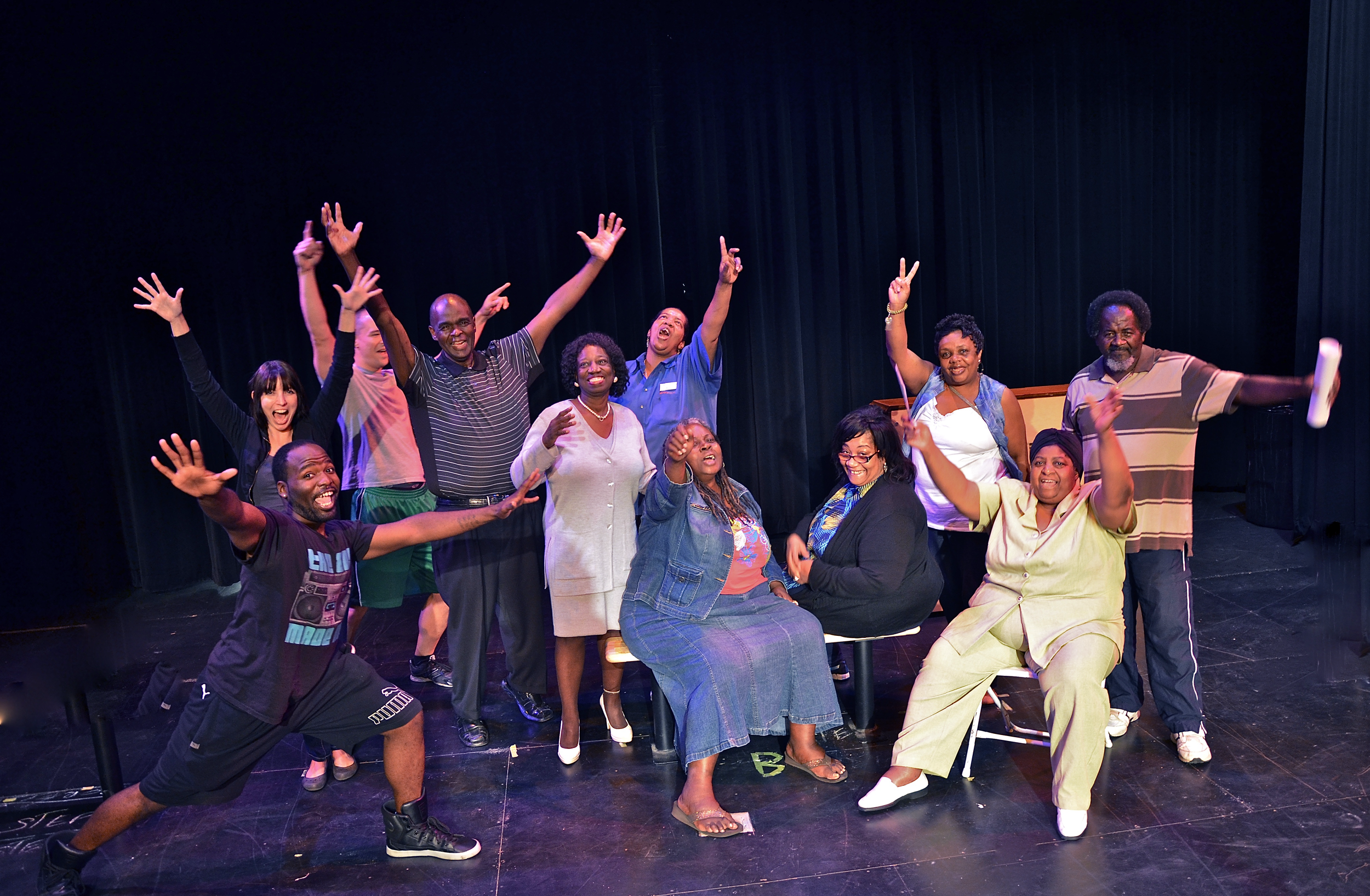 Group of diverse people smiling and posing excitedly on stage with black curtains in the background.