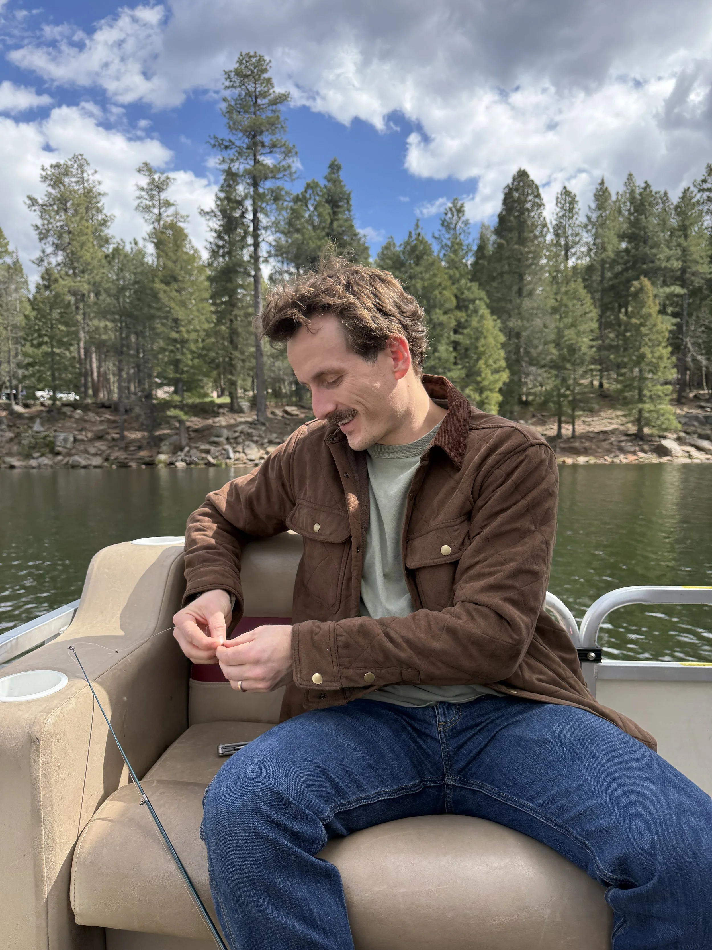 A man with brown curly hair and a mustache, wearing a brown jacket and blue jeans, sitting on a boat on a lake in the woods, smiling and looking down while holding something in his hands. The background shows tall green pine trees and a partly cloudy sky.