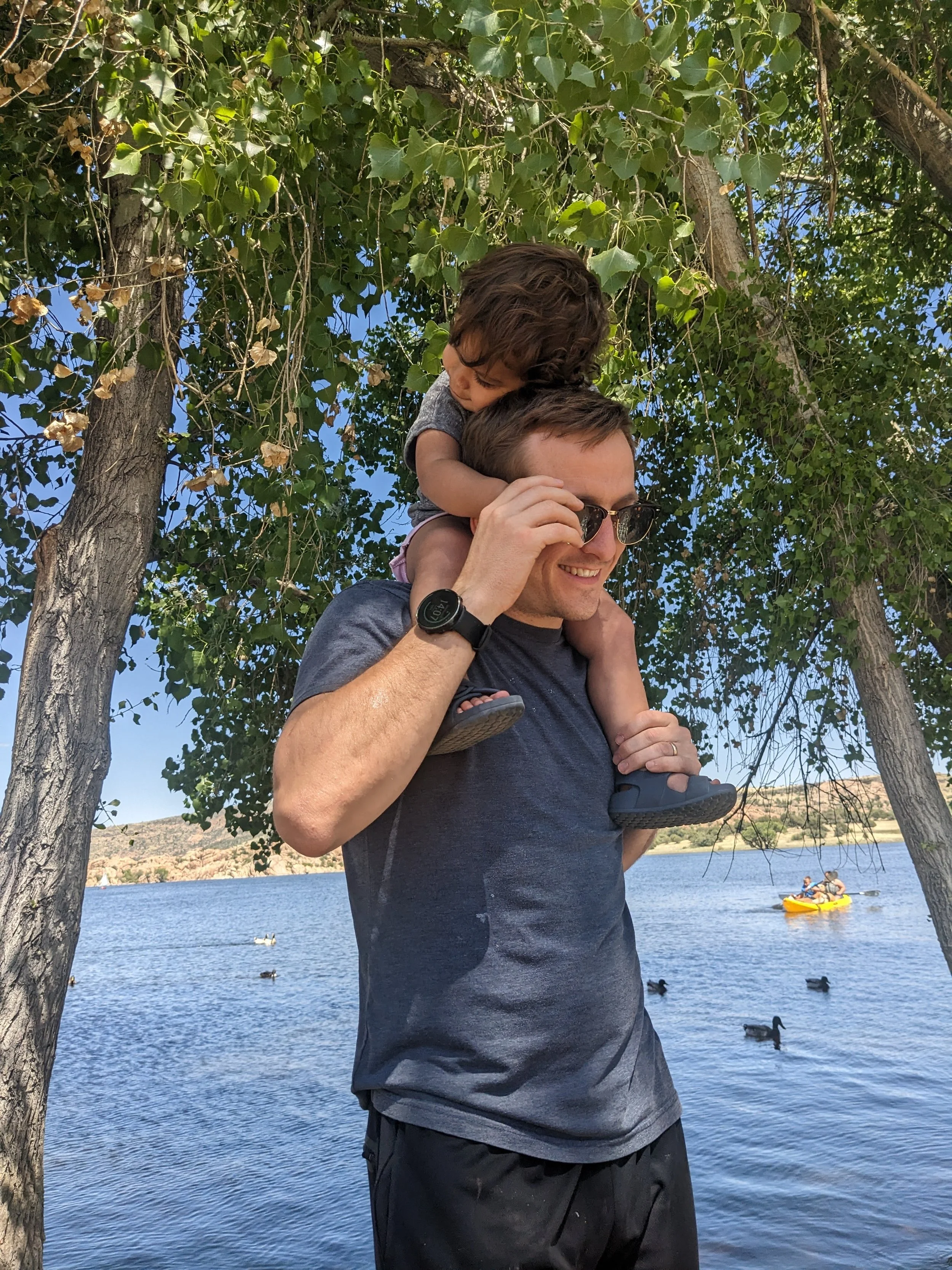 A man with short hair and glasses carrying his young daughter on his shoulders near a body of water, with ducks swimming and a small boat with two people in the background.