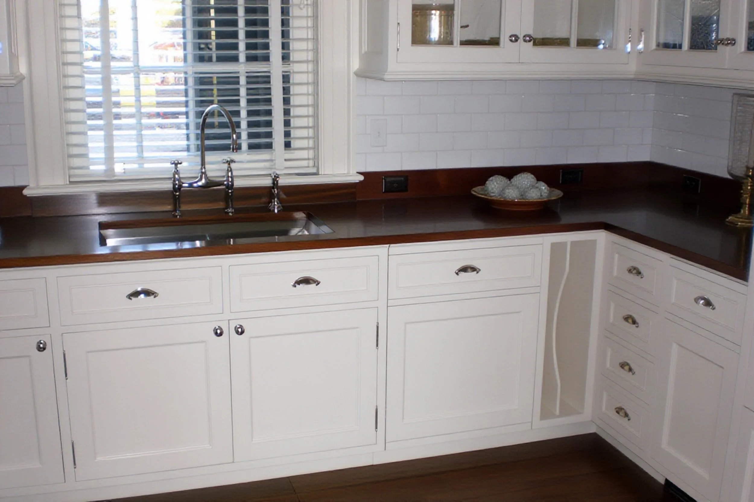 A kitchen with white custom cabinets, a dark wooden countertop, a stainless steel sink and a window with white blinds.