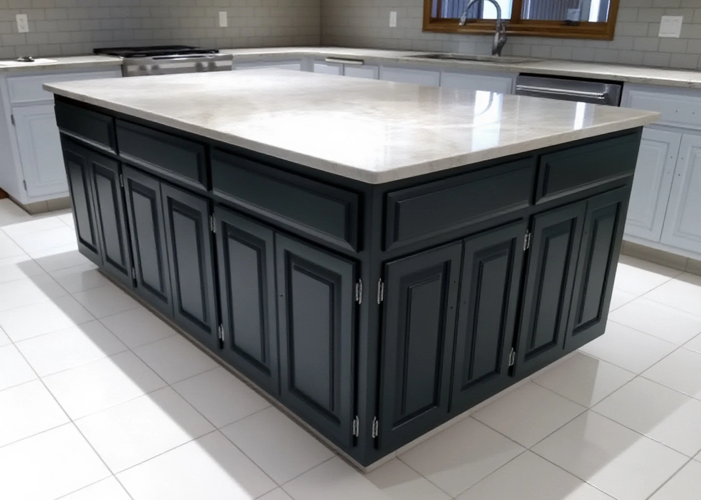 A kitchen island with a light-colored countertop and custom flat black cabinetry, surrounded by white cabinets, with a window and gray tiled wall in the background.