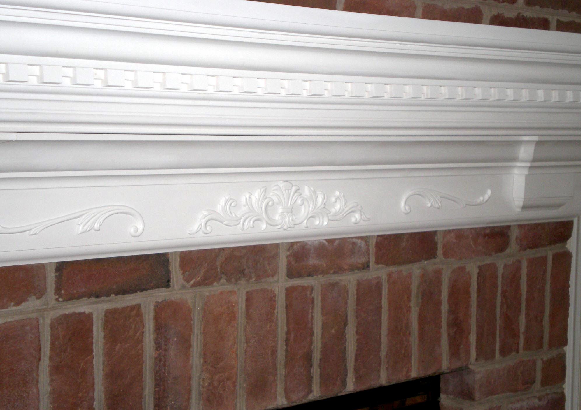 Close-up of a white, custom-carved decorative fireplace mantle with ornate detailing, mounted on a brick wall.
