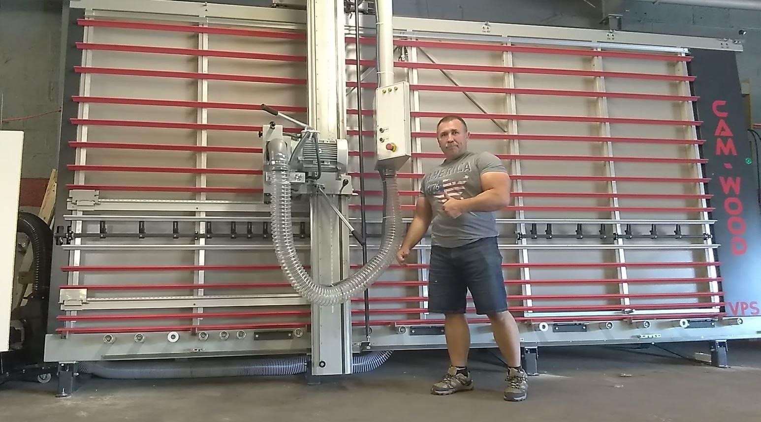 A man in a gray t-shirt and shorts standing next to a large panel saw in a cabinetry shop.