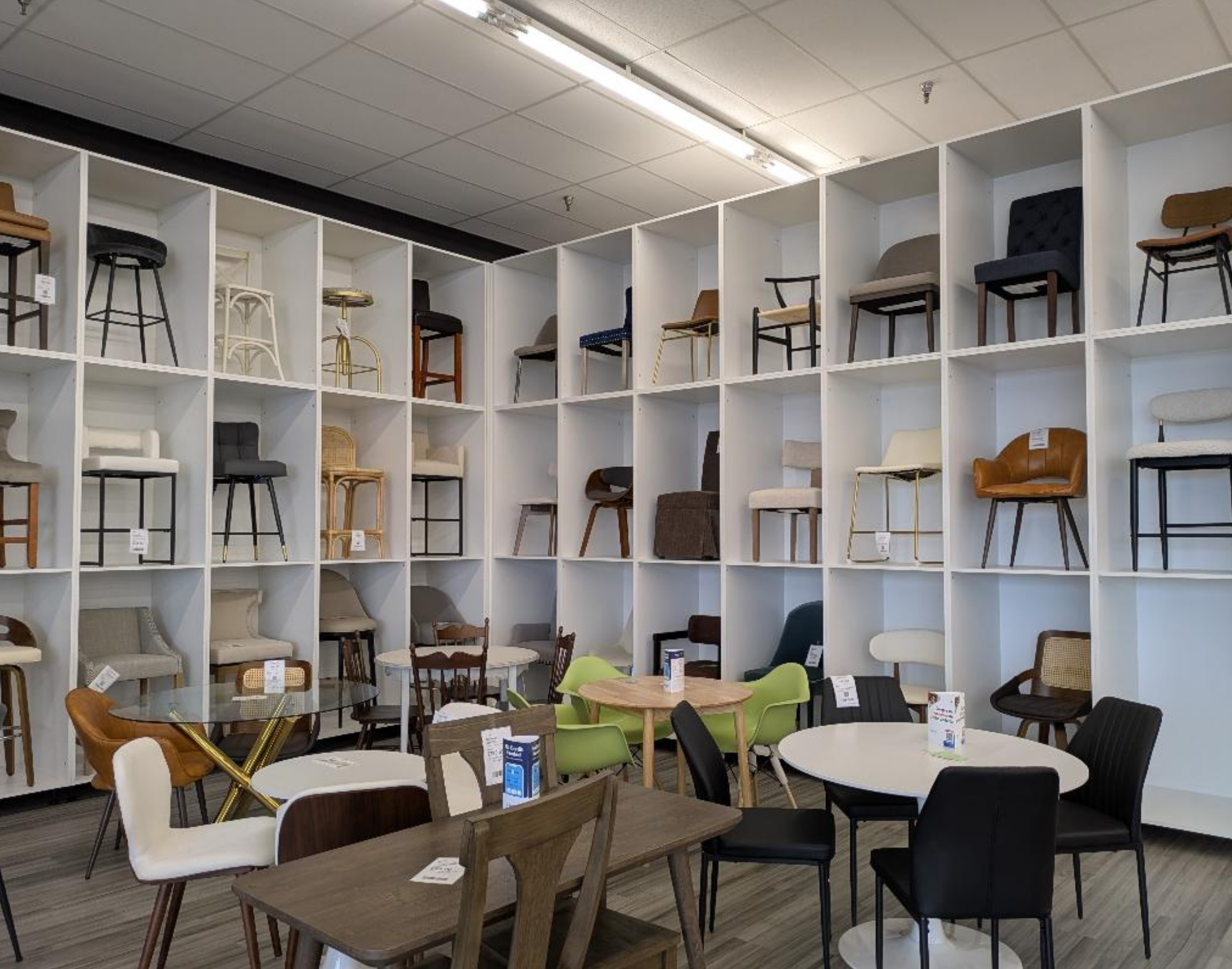 Store display of various chairs arranged on white, custom made white cubical shelves against a ceiling with fluorescent lighting, with tables and chairs set up in the foreground