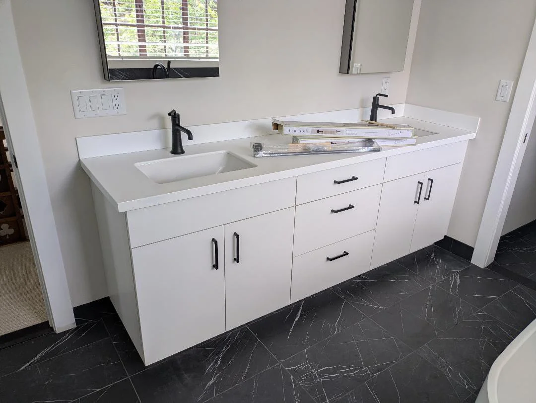 A custom made modern white bathroom vanity with two sinks, black faucets, multiple drawers, and cabinets, with a mirror above each sink, sitting a black marble floor.