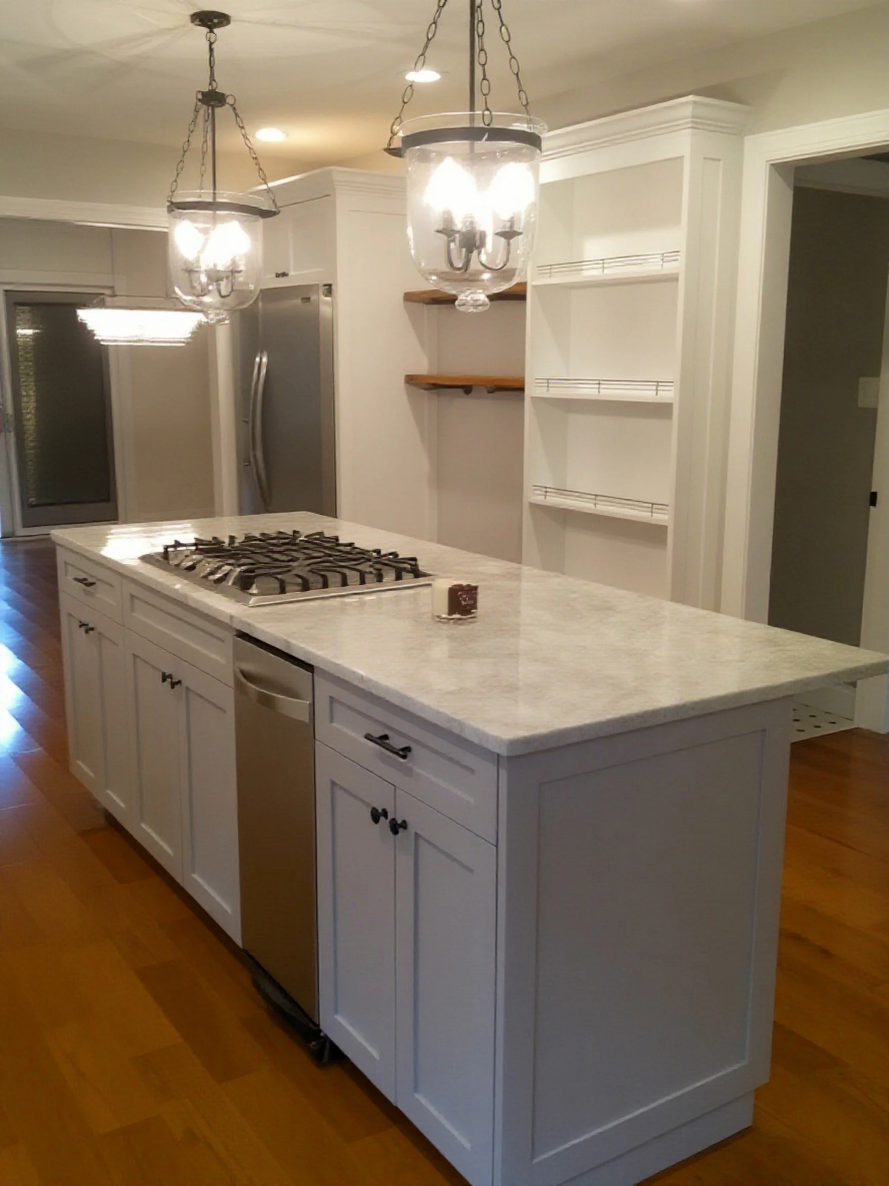 Kitchen island with a marble countertop, custom cabinetry, a gas stove, and black handles. Two hanging light fixtures with glass enclosures are above the island. In the background, stainless steel refrigerator and custom open shelving are visible.