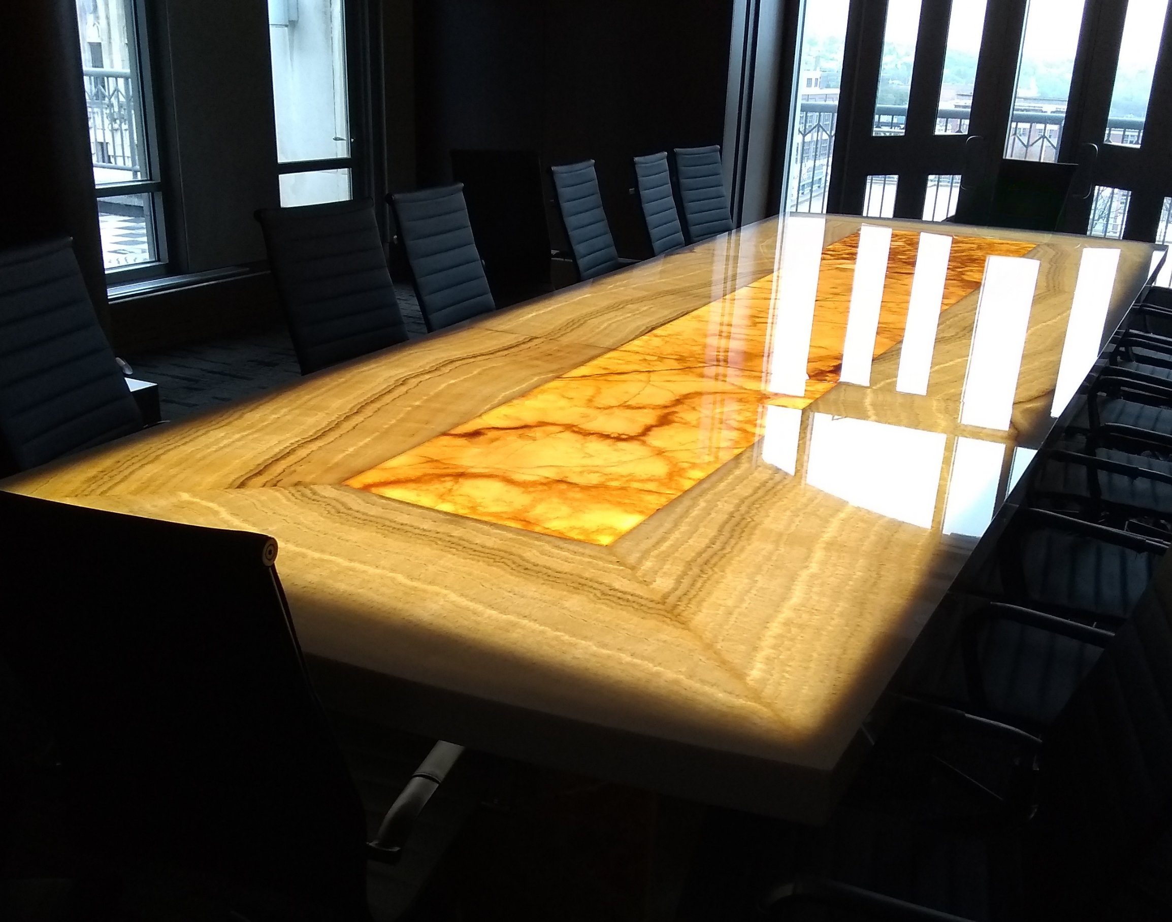 A conference room with a long, polished wooden table, custom made from scratch, surrounded by black leather office chairs, with windows and balcony doors allowing natural light.