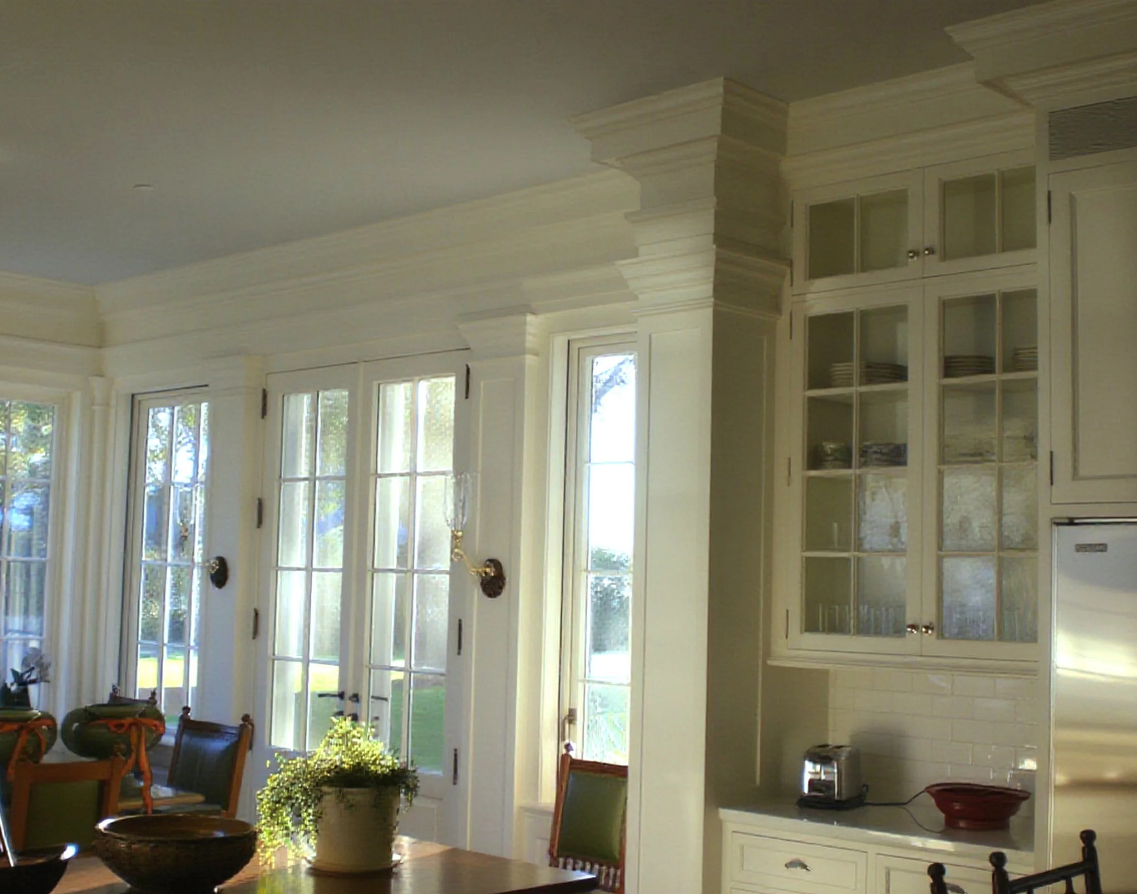 Interior view of a bright, spacious kitchen with large windows, white cabinets with glass doors, and a ceiling with custom crown molding. There are chairs and kitchen appliances visible.
