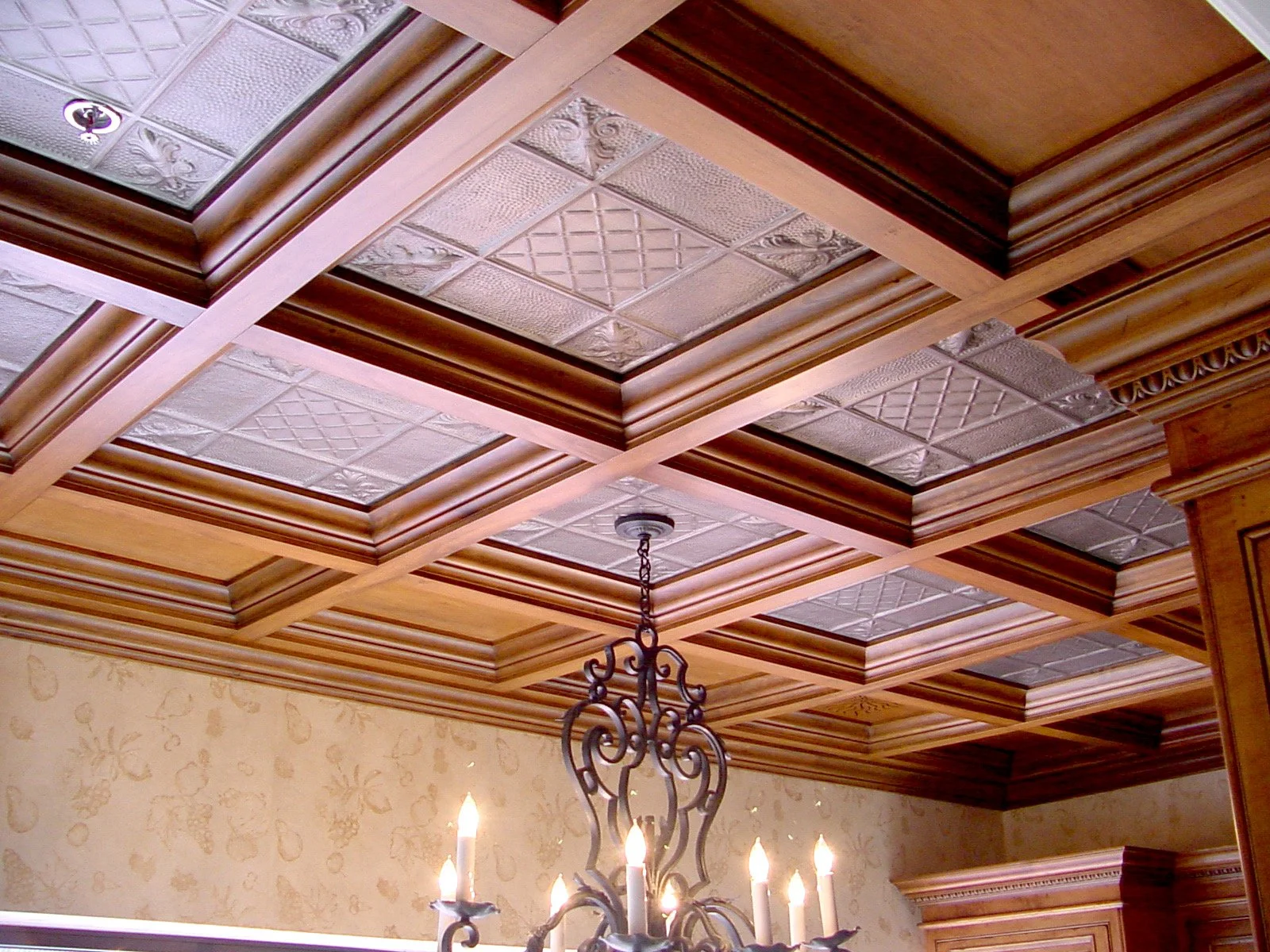 Custom coffered ceiling with wooden beams and ornate ceiling tiles, chandelier with lit candles hanging from the ceiling, and wallpapered wall with a decorative pattern.