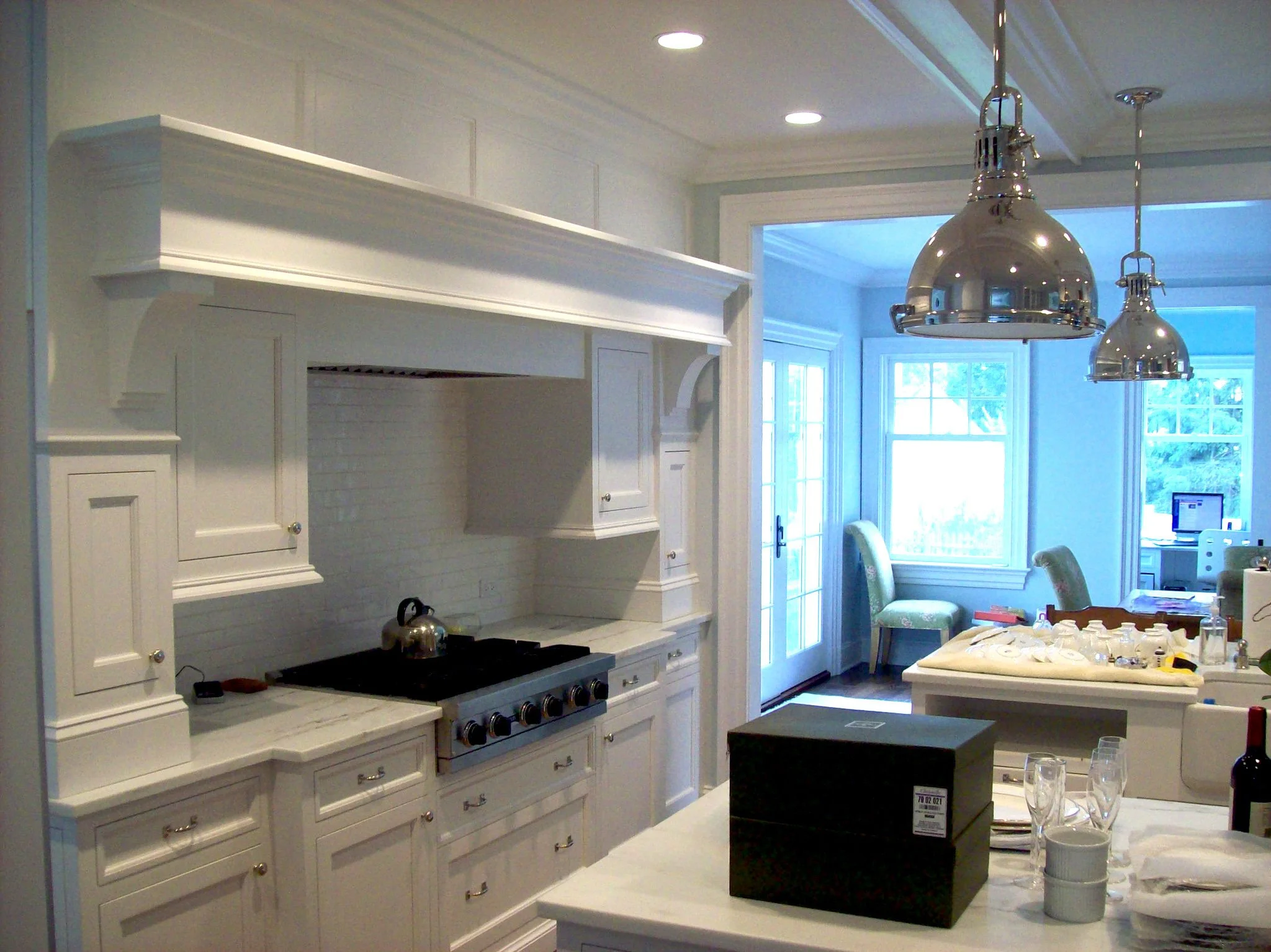White kitchen with custom white cabinetry, stainless steel range with black stovetop, and two large silver pendant lights hanging above a kitchen island. Large windows with sliding glass door in the background, with a view of outdoor greenery.