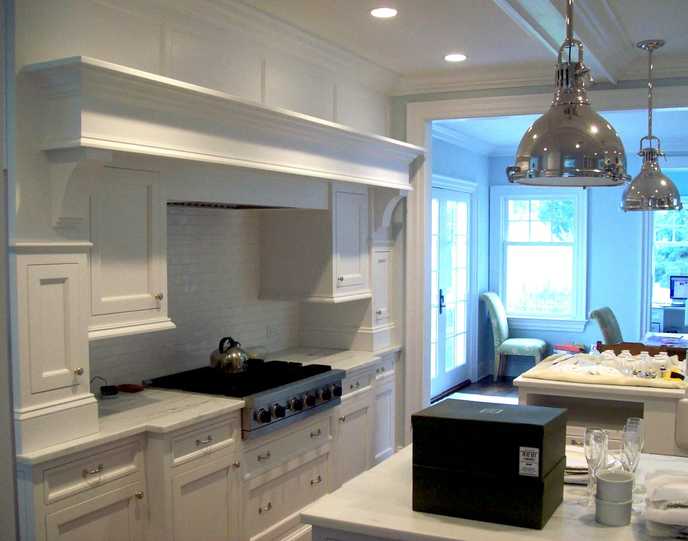White kitchen with custom cabinetry, stainless steel range with black stovetop, and two large silver pendant lights hanging above a kitchen island alongside custom carved moldings. Large windows with sliding glass door in the background, with a view 