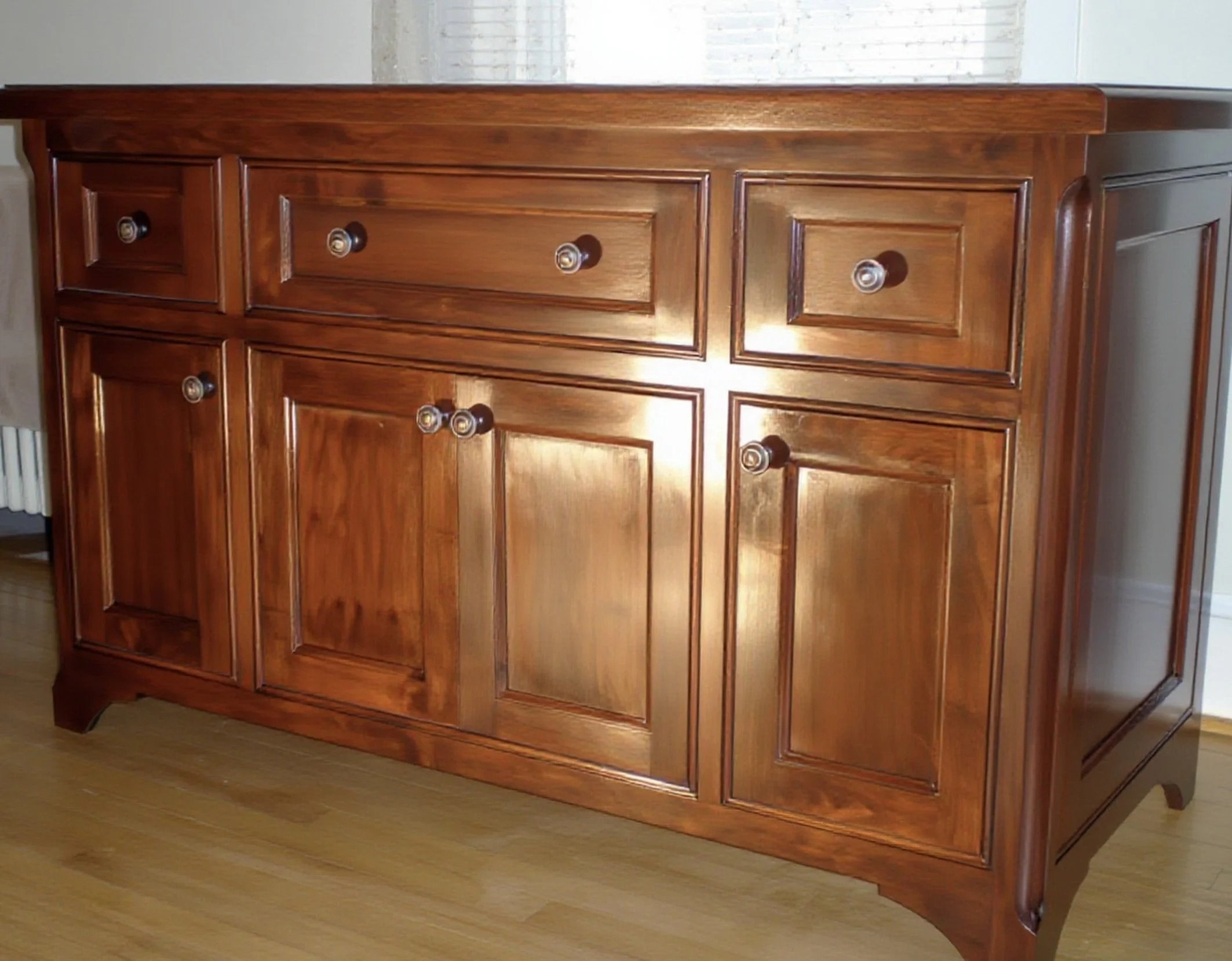 A made-to-order wooden cabinet with six drawers and round knobs, placed on a hardwood floor near a white wall with a window in the background.