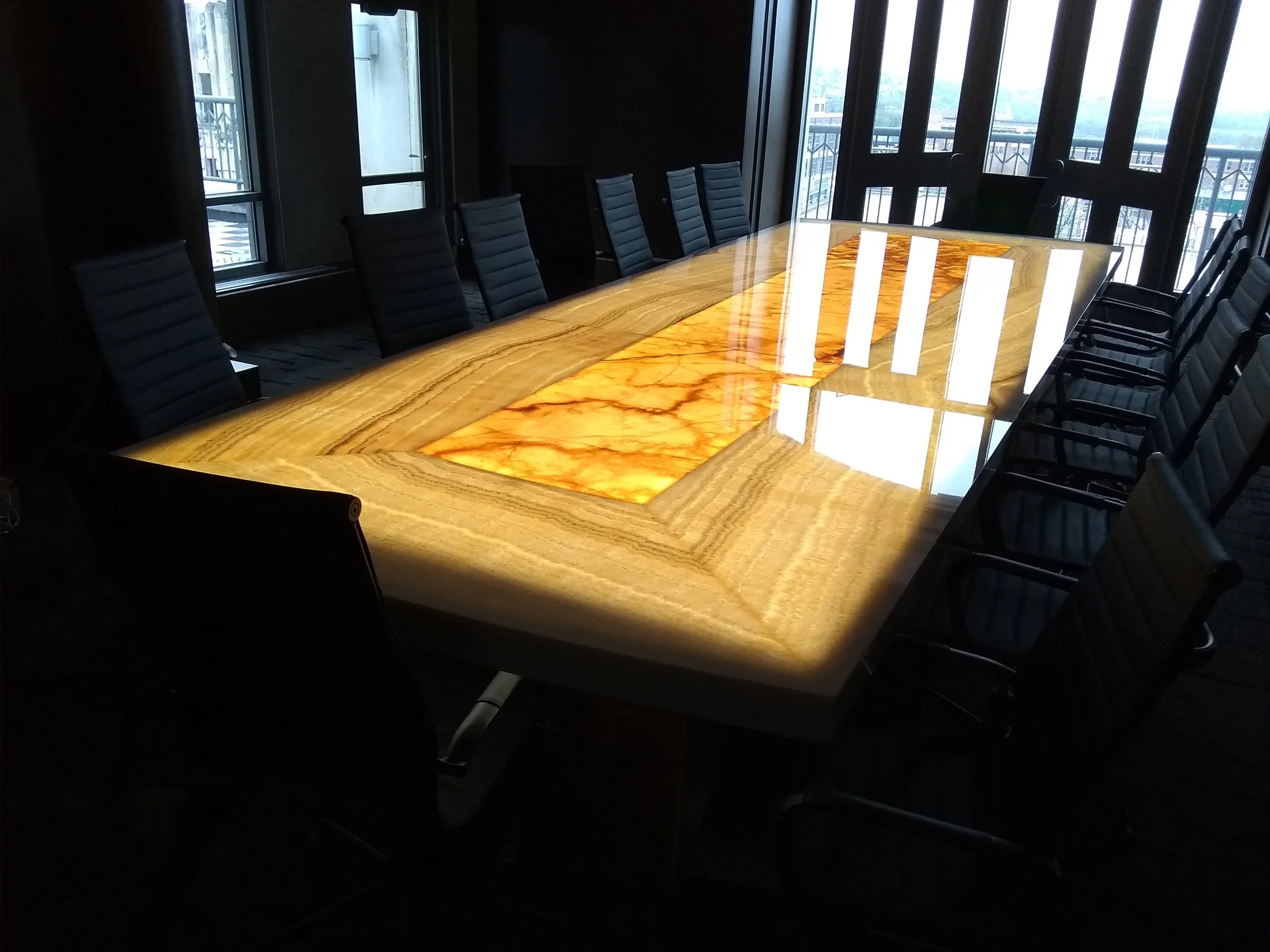 A conference room with a large custom-made polished wooden table, surrounded by black leather chairs, and large windows revealing a cityscape outdoors.