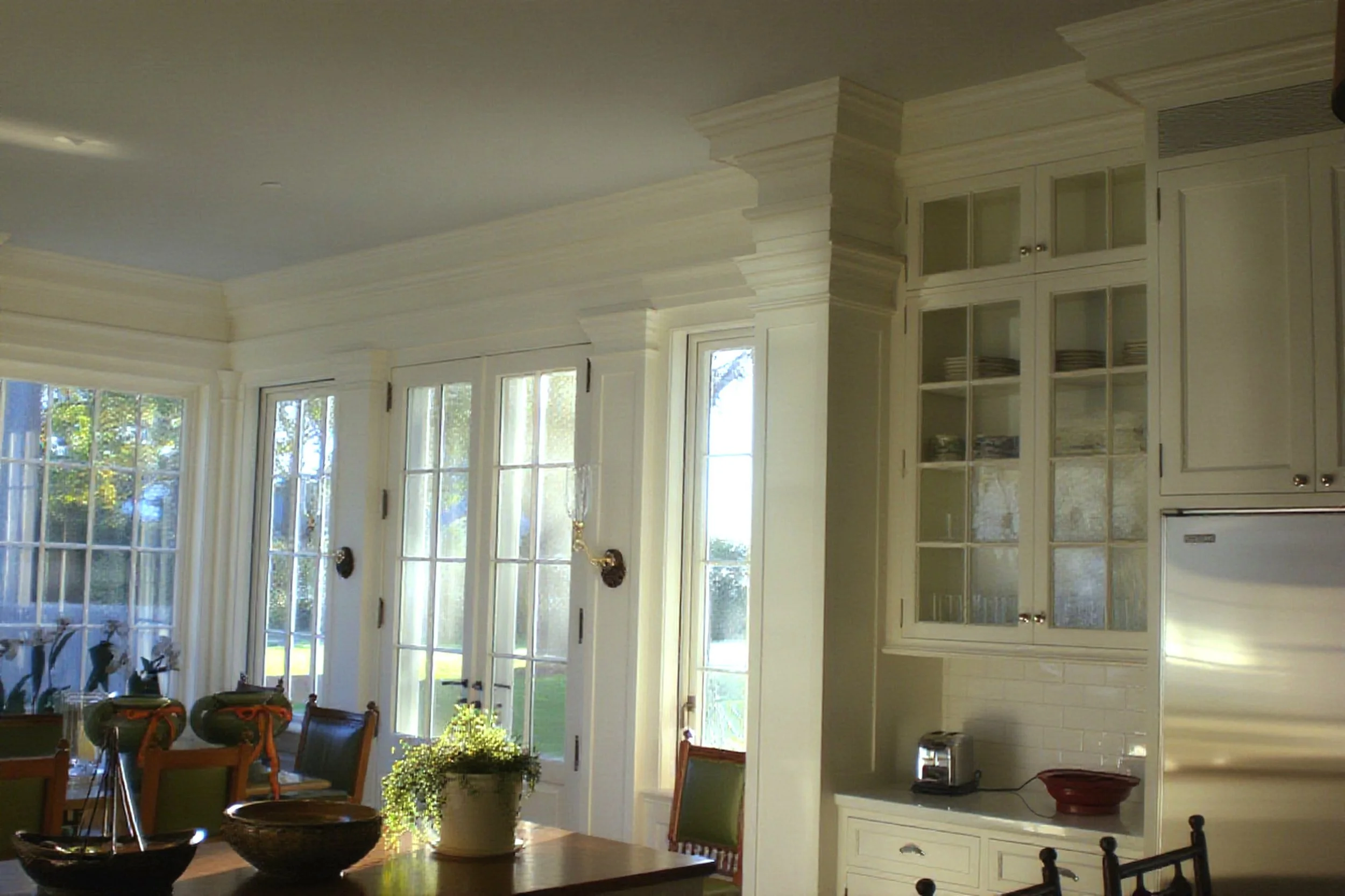 A bright kitchen with large windows letting in natural light, white custom cabinets with glass-paneled doors, a wooden dining table with chairs, and decorative bowls and plants on the table.