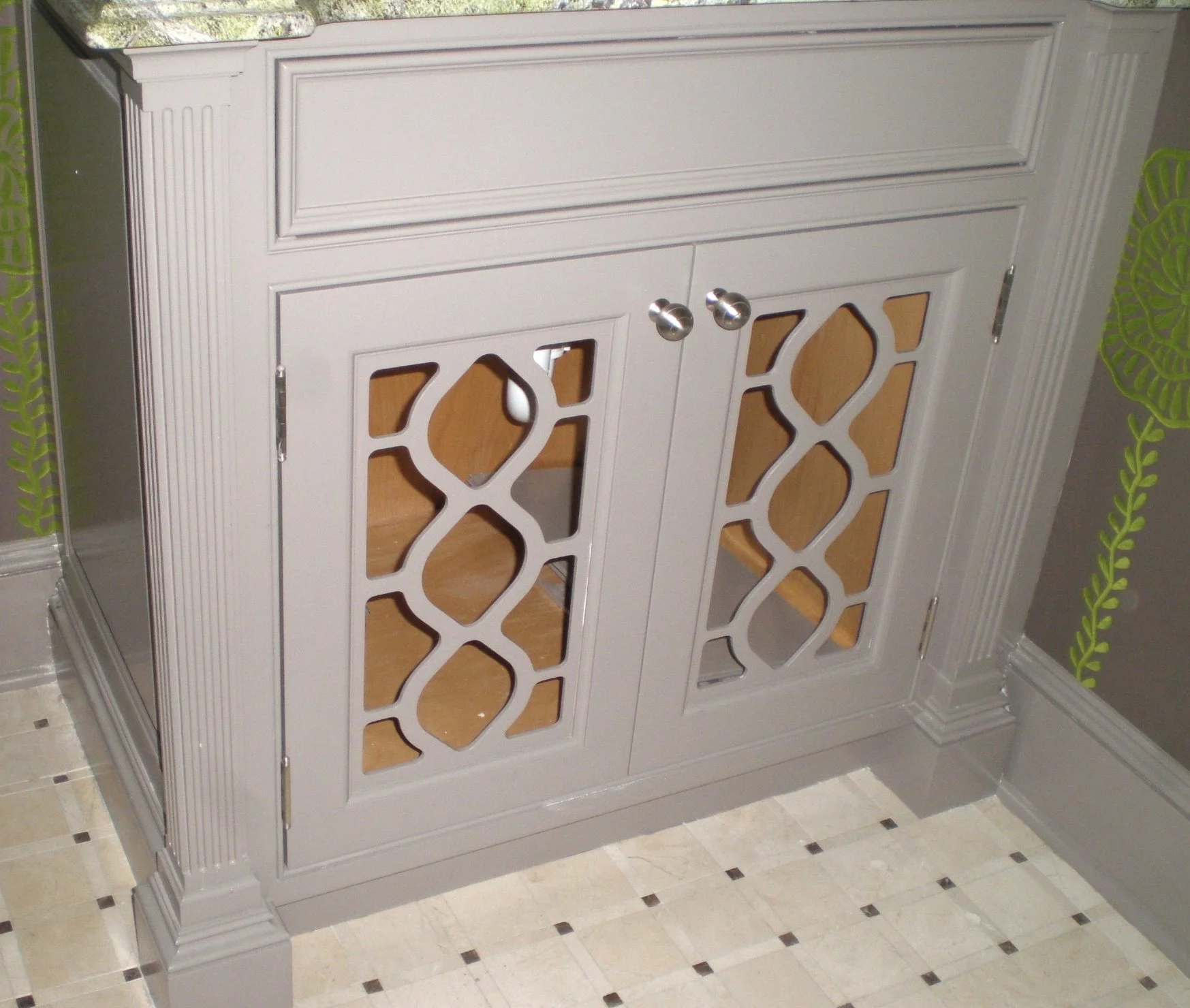 A beige cabinet with decoratively cut doors and silver knobs in a room with a checkered floor and green patterned wall.