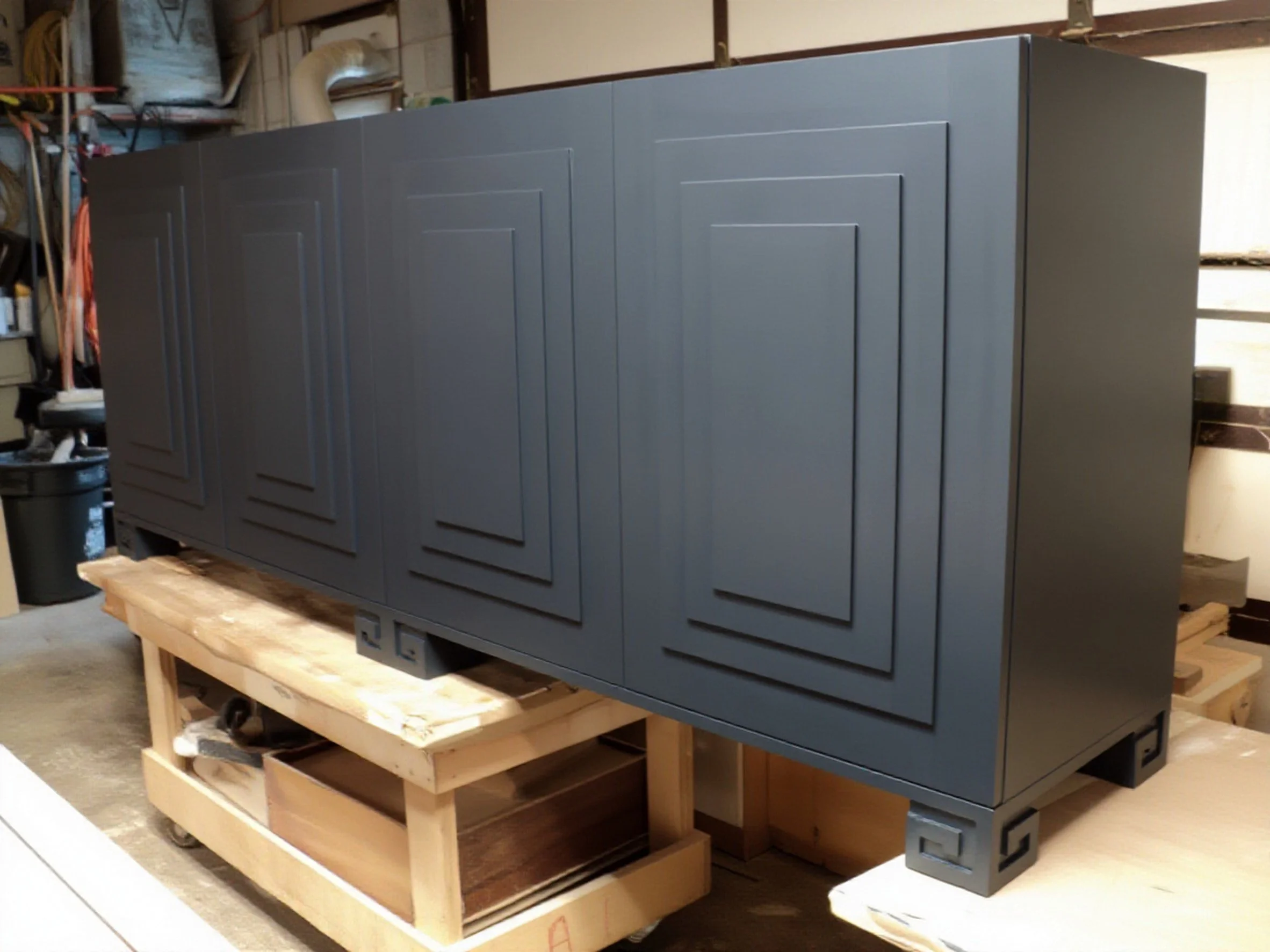 A handcrafted large, matte black painted cabinet with decorative molding on the doors, sitting on a wooden bench in a workshop. 